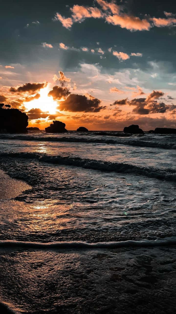 Coastal Sunset 桌布 壁紙 — 夕陽將天空染成火熱的橙色和深藍色，海浪拍打著寧靜海岸線上的黑色岩石。