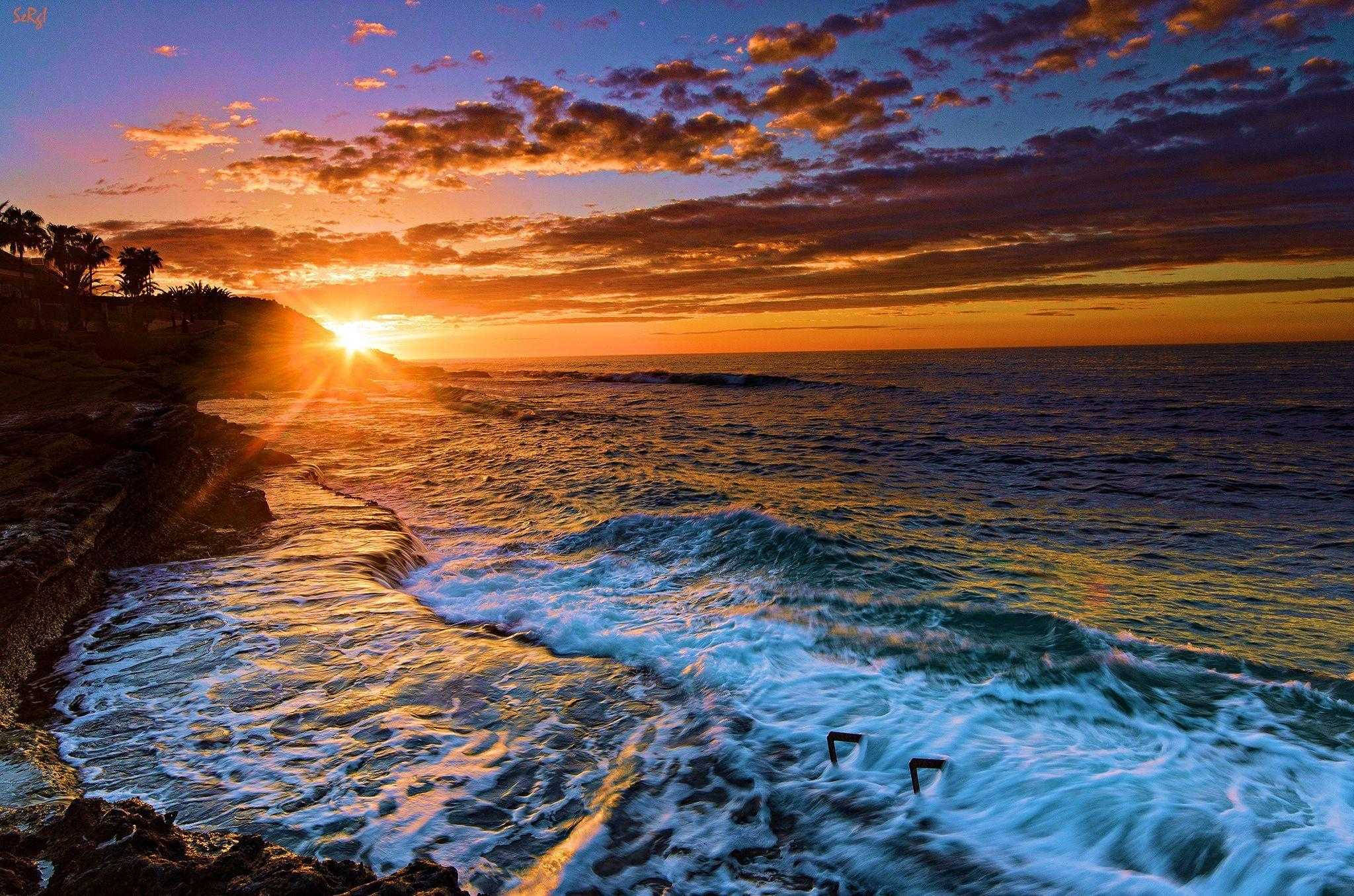 Sunset Beach 桌布 壁紙 — 夕陽將天空染成火熱的橙色和紫色，海浪拍打岩石海岸，棕櫚樹在懸崖上形成剪影。