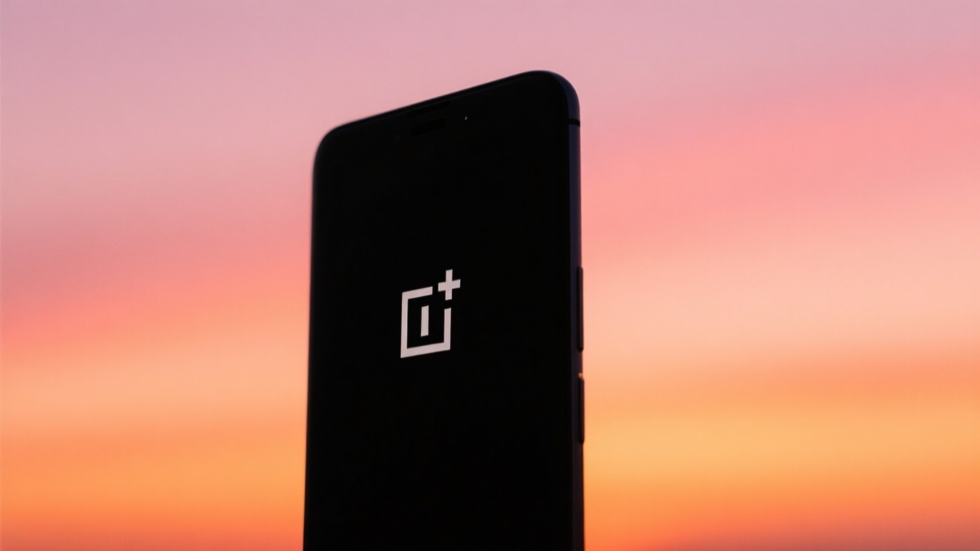 Sunset Phone Silhouette 壁紙 — 一部黑色 OnePlus 智能手機直立於充滿活力的日落天空前，混合了粉紅、橙和蜜桃色調。