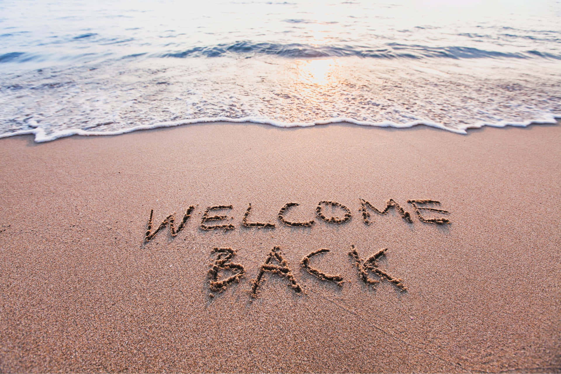  壁紙 — 夕陽下的海浪輕輕拍打沙灘，沙上寫著逐漸褪色的「WELCOME BACK」。