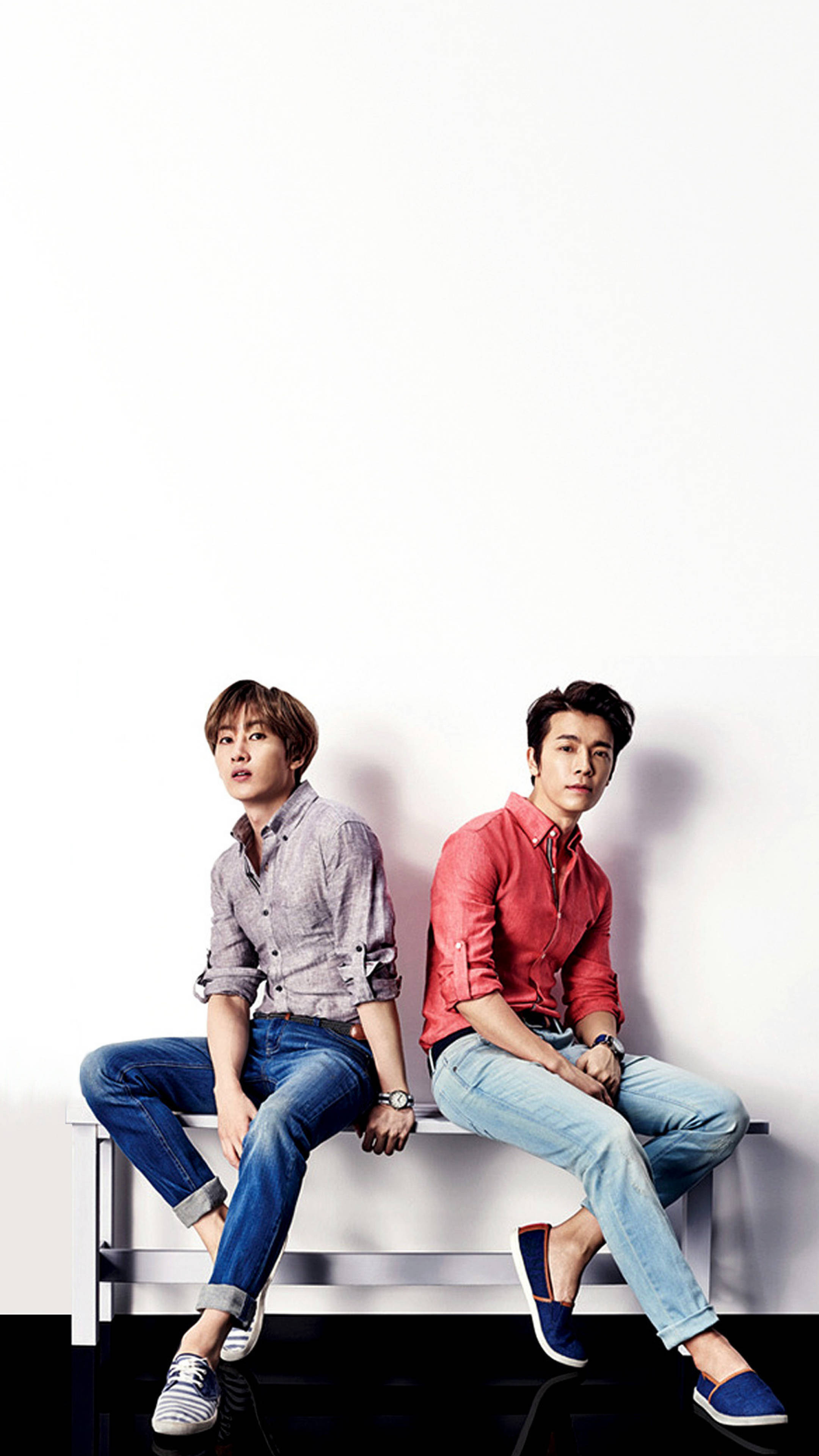 Super Junior 鄭允浩與東海 壁紙 — 兩位時尚年輕男子並排坐在簡約長椅上，背景是純白牆壁，身穿休閒牛仔褲和色彩繽紛的襯衫。