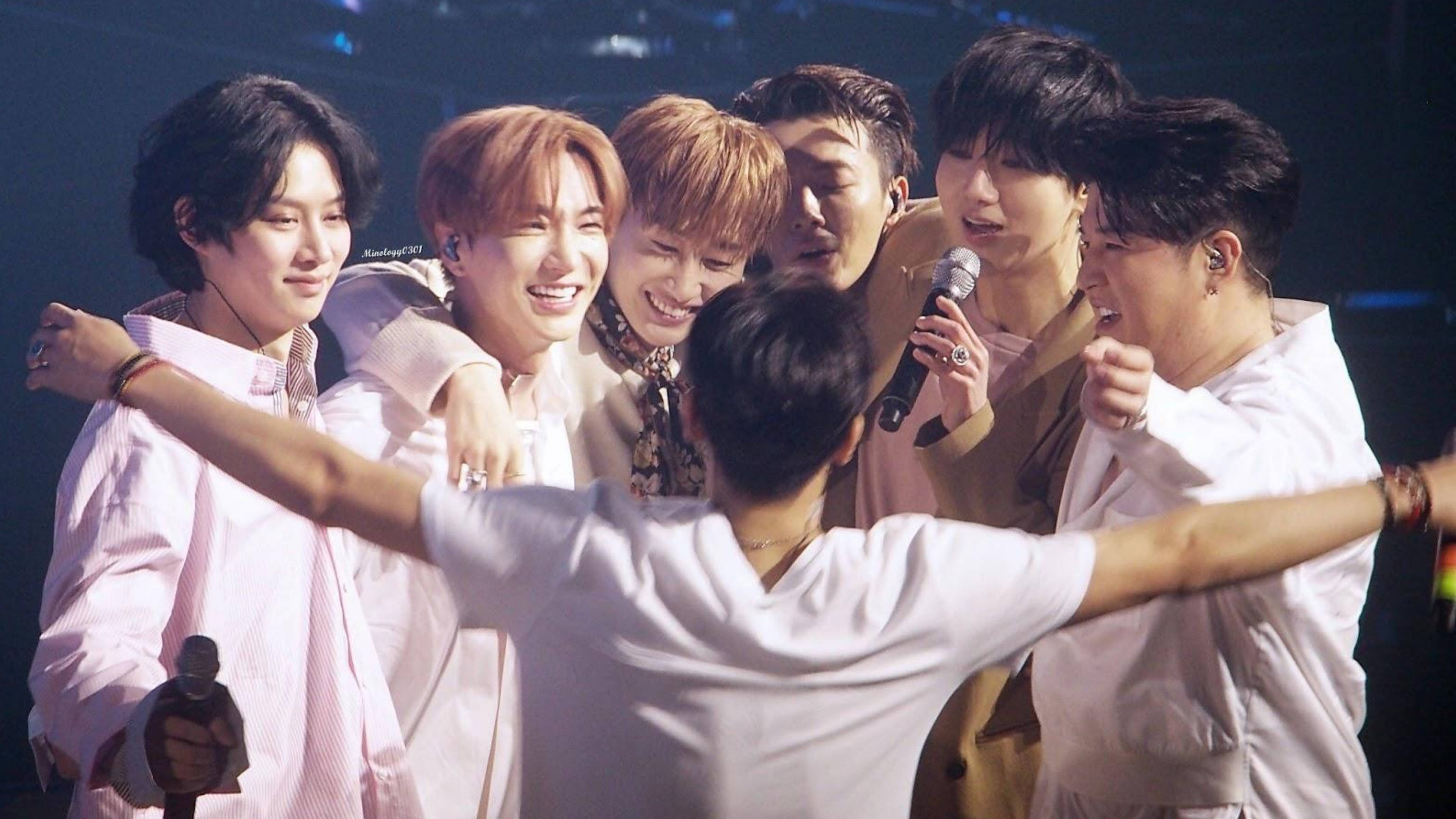 Super Junior集體擁抱 壁紙 — 七位面帶微笑的男表演者身穿淺色襯衫，在燈光昏暗的舞台上緊密地聚在一起，其中一人手持麥克風，另一人拿著一個小獎盃。