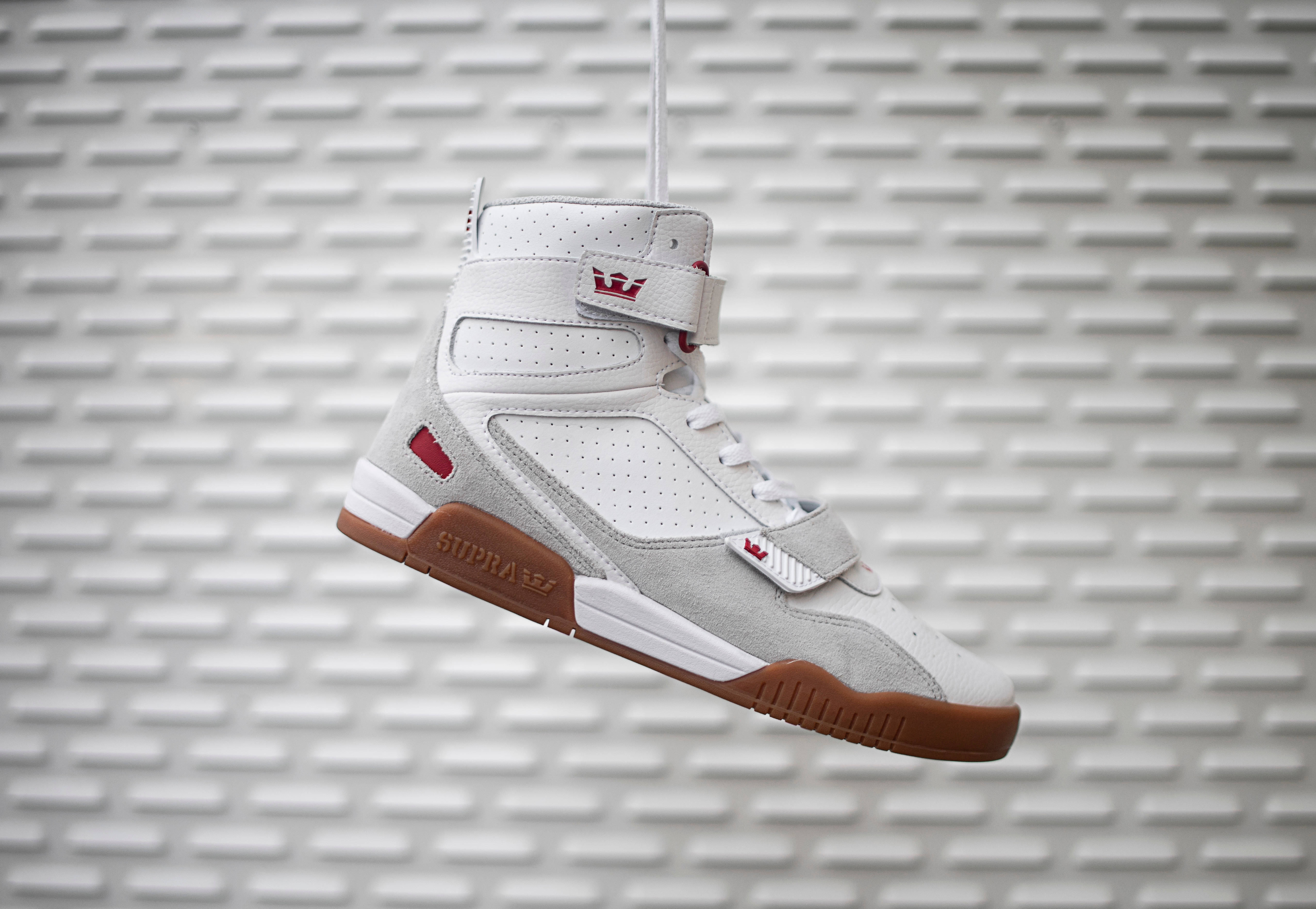 Supra Breaker高筒籃球鞋 壁紙 — 一雙帶有紅色點綴和棕色鞋底的白色高筒運動鞋懸掛在帶有紋理的灰色磚牆前。