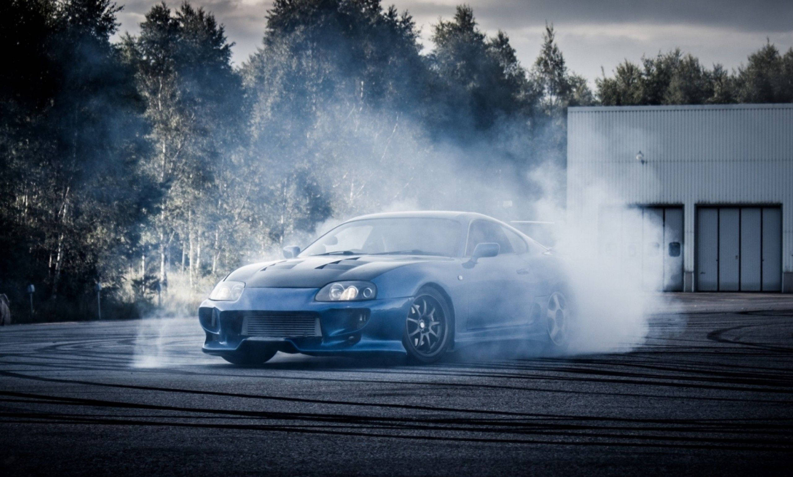 煙熏藍豐田Supra 壁紙 — 一輛藍色跑車在倉庫和樹木旁的瀝青場地上，在煙霧中飄移。