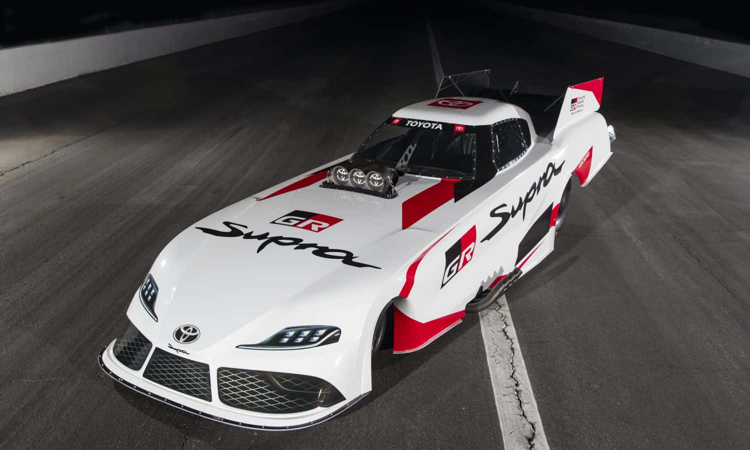 搞笑豐田Supra賽車圖 壁紙 — 一輛白色和紅色的豐田Supra賽車帶有'GR Supra'品牌標誌，在明亮的聚光燈下停泊在黑暗的賽道上。