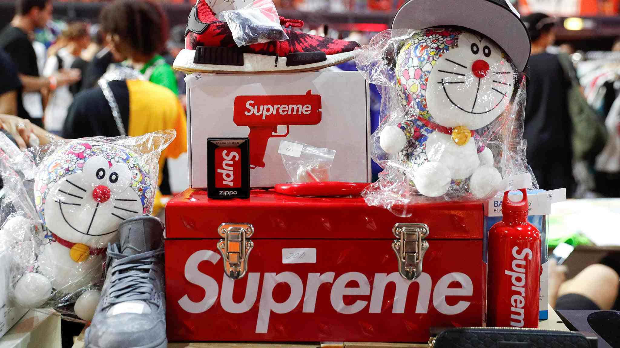 以 Supreme 穿搭時尚 壁紙 — 一個紅色Supreme行李箱展示了一系列品牌商品，包括多啦A夢毛公仔、運動鞋和Zippo打火機，在一個擁擠的快閃店內。