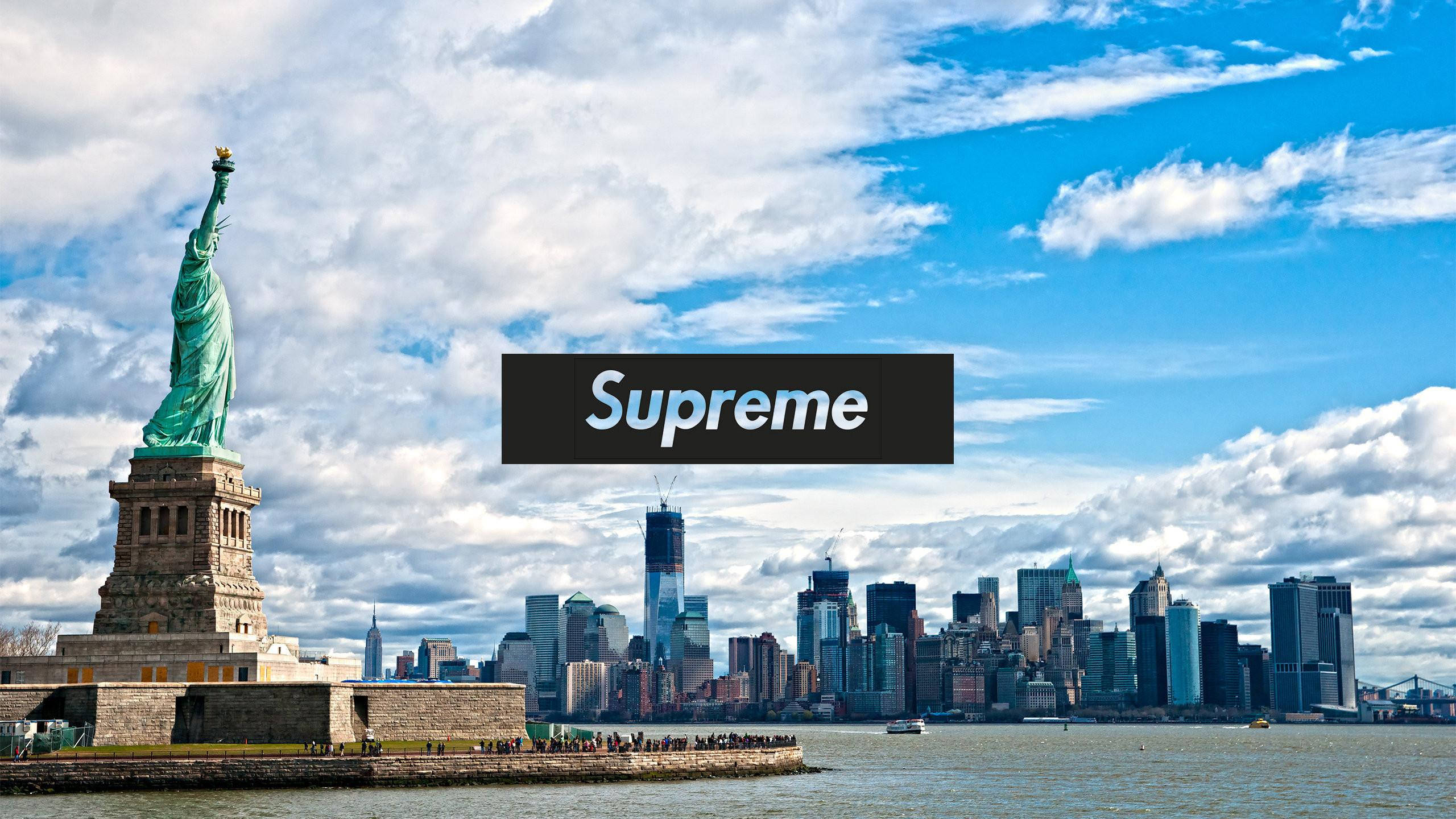酷炫 Supreme 自由女神像 壁紙 — 自由女神像在明亮的藍天和蓬鬆的白雲下，高聳於紐約市天際線旁。