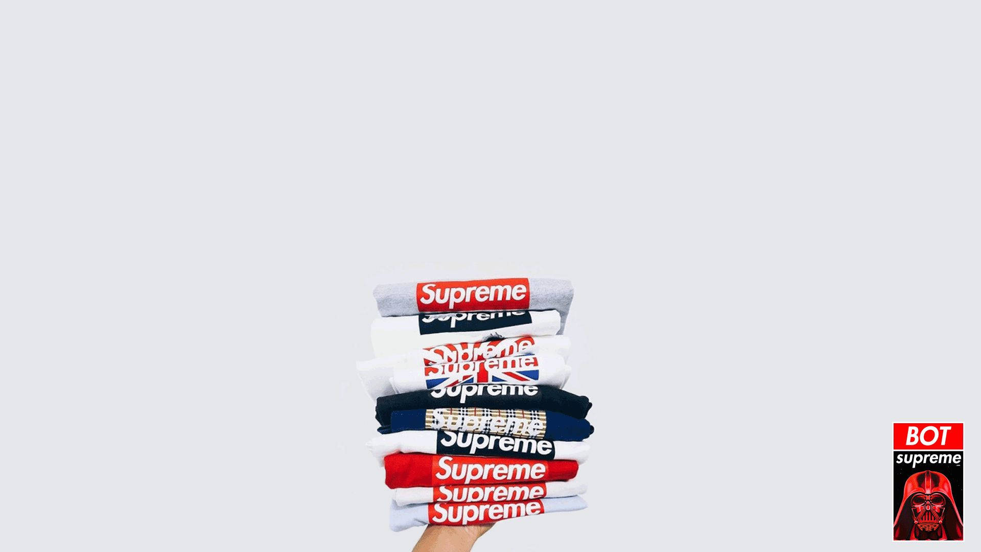 Supreme襯衫堆疊 壁紙 — 一隻手拿著一疊Supreme品牌T恤，背景是純淺灰色，角落有一個小小的Darth Vader標誌。