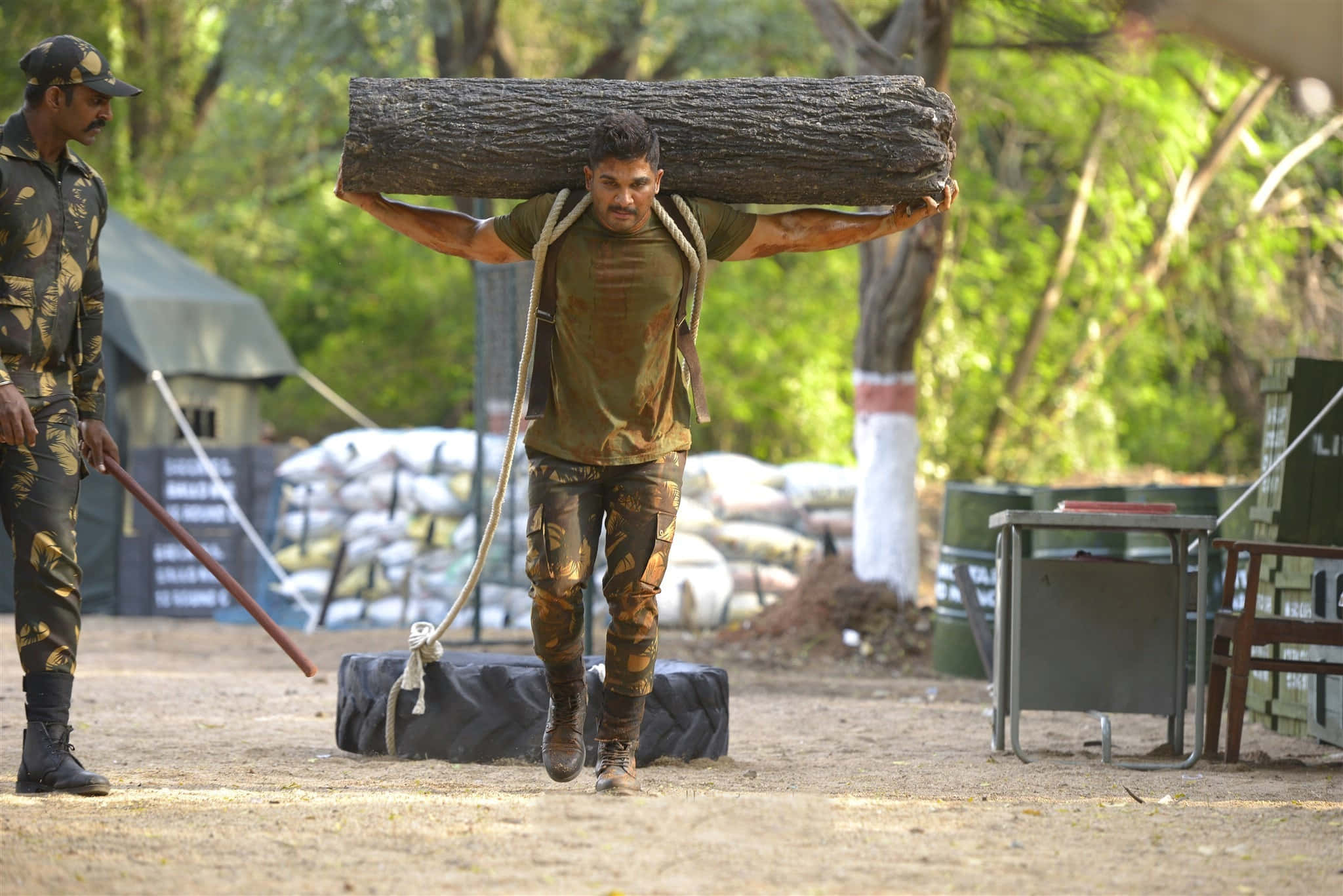 「Surya，堅定的戰士」——電影《Surya The Soldier》劇照，Allu Arjun飾演 壁紙 — 一名士兵在泥濘的戶外訓練場上扛著沉重的圓木行走，另一名士兵在旁觀看。