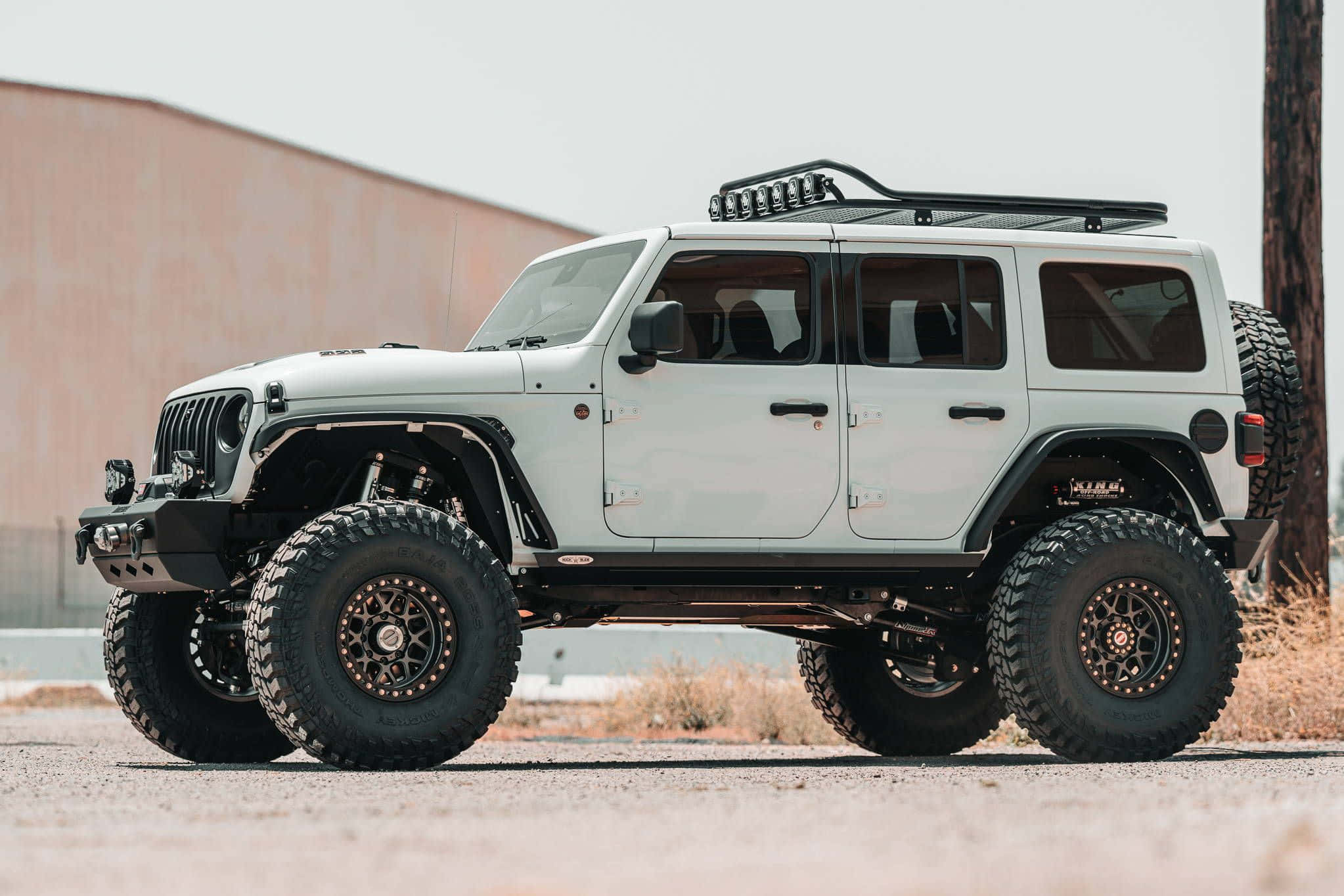 越野冒險：強悍SUV征服崎嶇地形 壁紙 — 一輛白色升高嘅Jeep Wrangler，配備超大輪胎同車頂行李架，停泊喺碎石上，附近有木柱同米色牆。