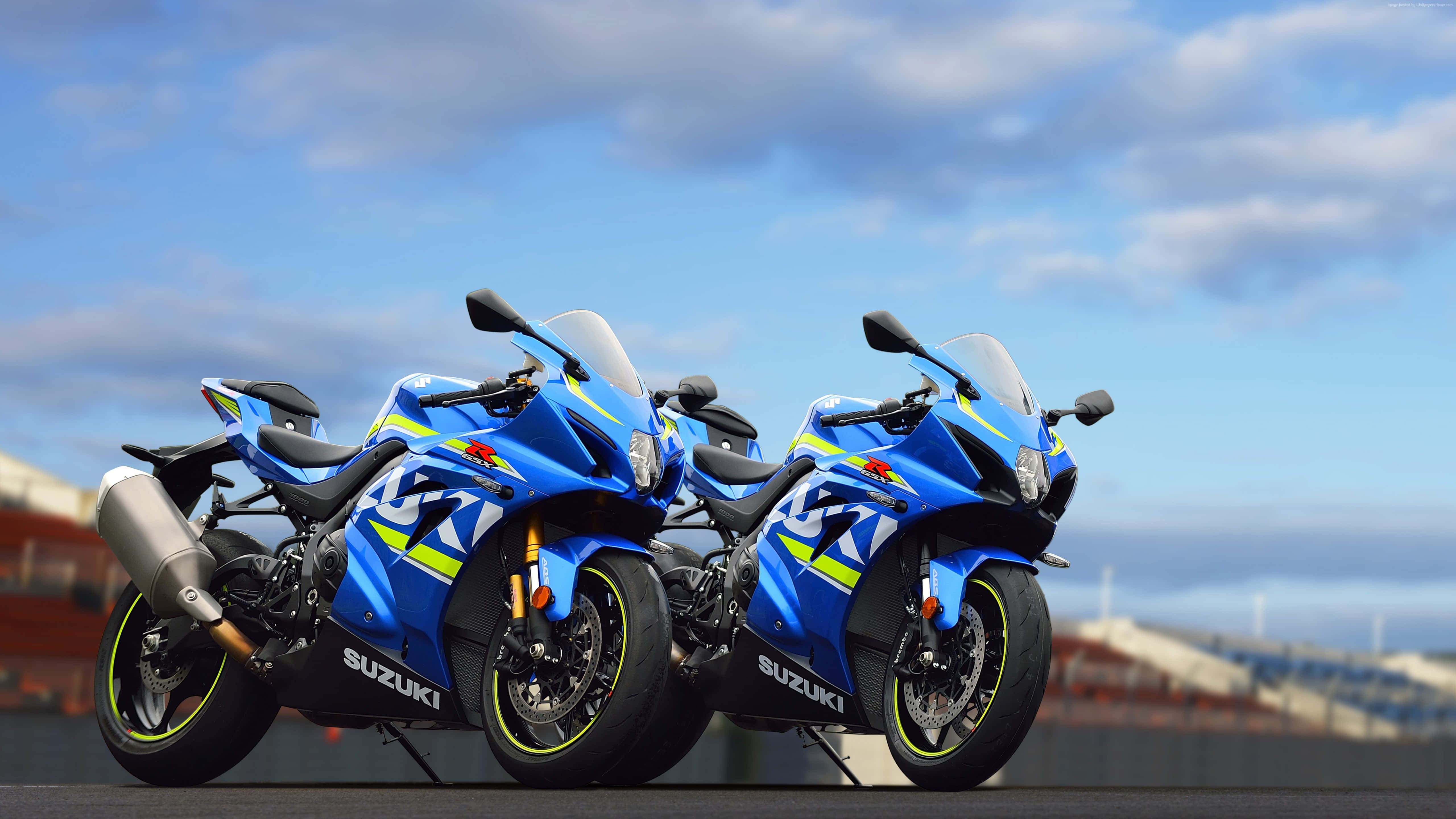 Suzuki GSX R1000 最佳機車背景 壁紙 — 兩輛藍色鈴木運動電單車帶有霓虹燈飾，並排停泊在陰天下的賽車場上。