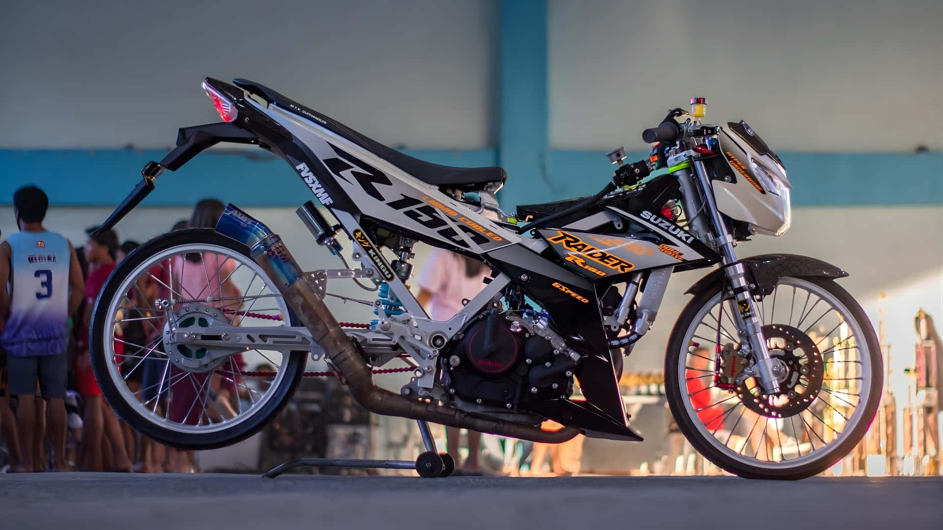  壁紙 — 一架時尚的黑白 Suzuki 越野電單車帶有橙色點綴，在黃昏時立於支架上，背景有模糊的觀眾。