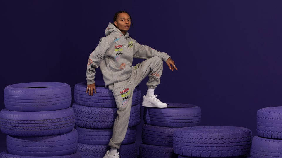 Swae Lee 坐於紫色輪胎上 壁紙 — 一個人穿著塗鴉覆蓋的灰色連帽衫和運動褲，自信地擺姿勢在充滿活力的紫色輪胎堆上，背景是深紫色。