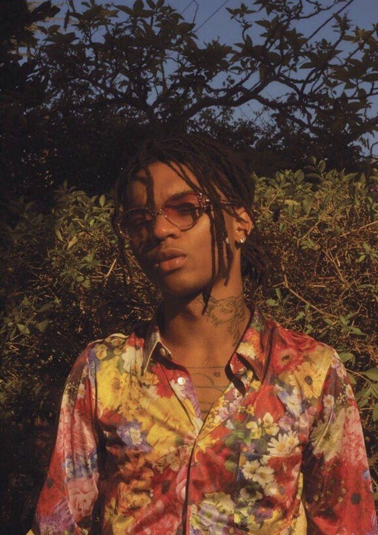 Swae Lee 身穿花卉襯衫 壁紙 — 一位戴著眼鏡、留著辮子頭的年輕男子，在金色時光的光線下，以茂密的樹葉為背景，穿著一件鮮豔的花襯衫。