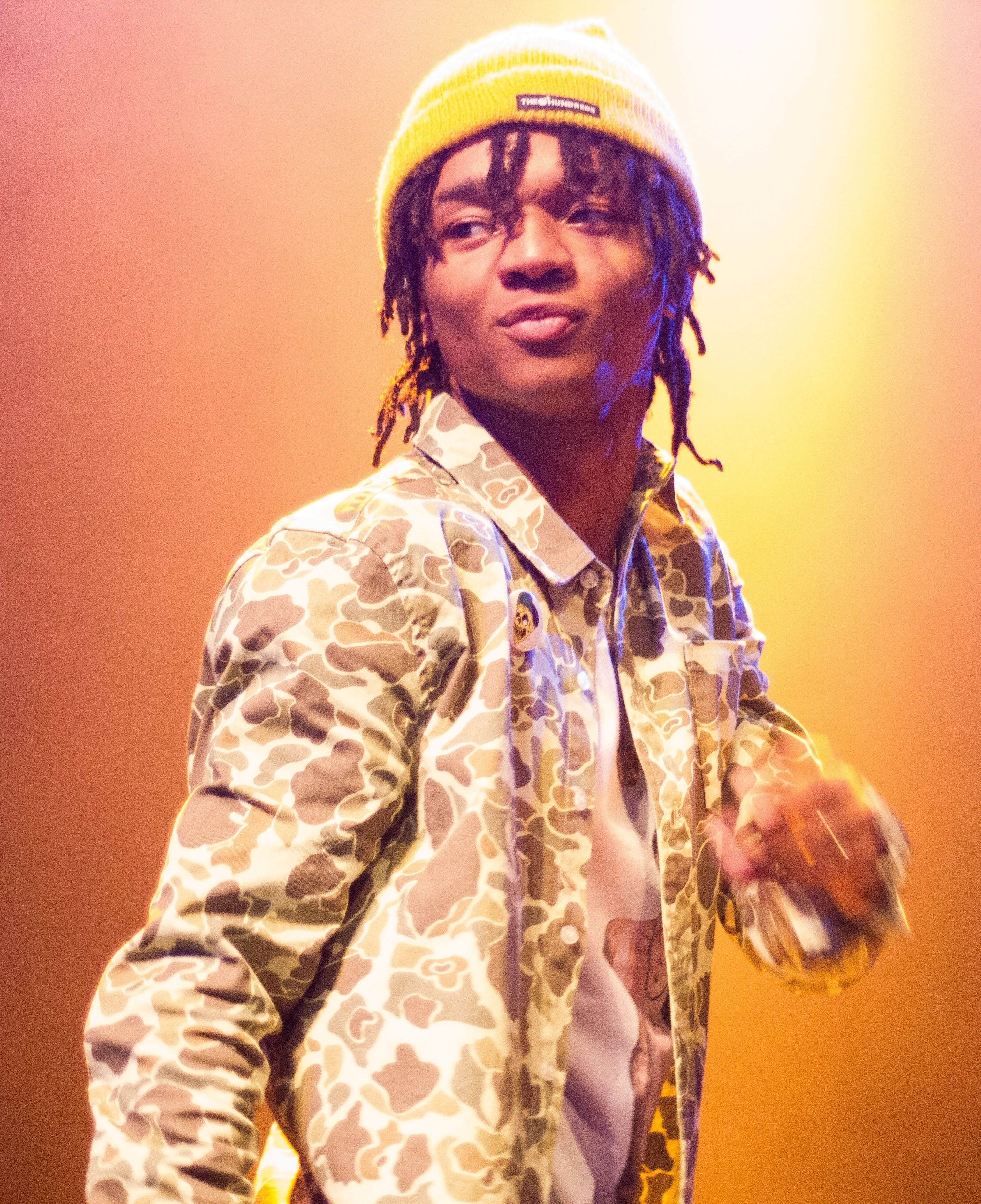 Swae Lee 在光線中 壁紙 — 一位年輕男子留著辮子頭，戴著黃色無邊帽，身穿迷彩外套，在溫暖的橙黃燈光下於舞台上表演。