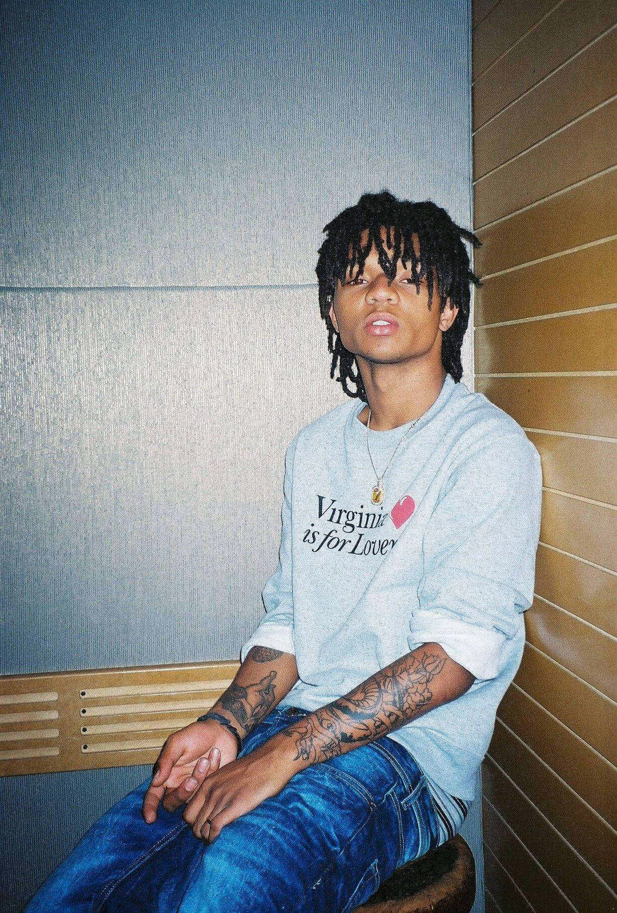Swae Lee 在攤位前 壁紙 — 一位留著辮子頭和紋身的年輕男子，隨意地坐在木長椅上，背靠有紋理的牆壁，身穿維珍尼亞運動衫。