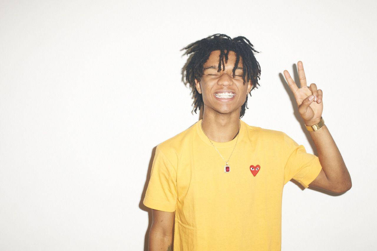 Swae Lee 身穿黃色襯衫 壁紙 — 一位留著辮子頭的年輕男子對著純白牆壁微笑並做出和平手勢。