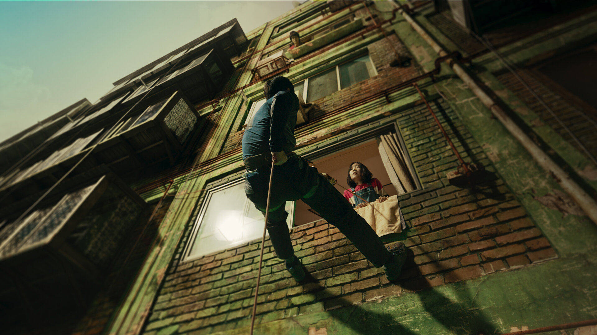 《Sweet Home》Netflix 韓劇：綠頂公寓劇照 壁紙 — 一個人攀爬一座風化的綠色磚建築，而一個孩子從下方的窗戶觀看。