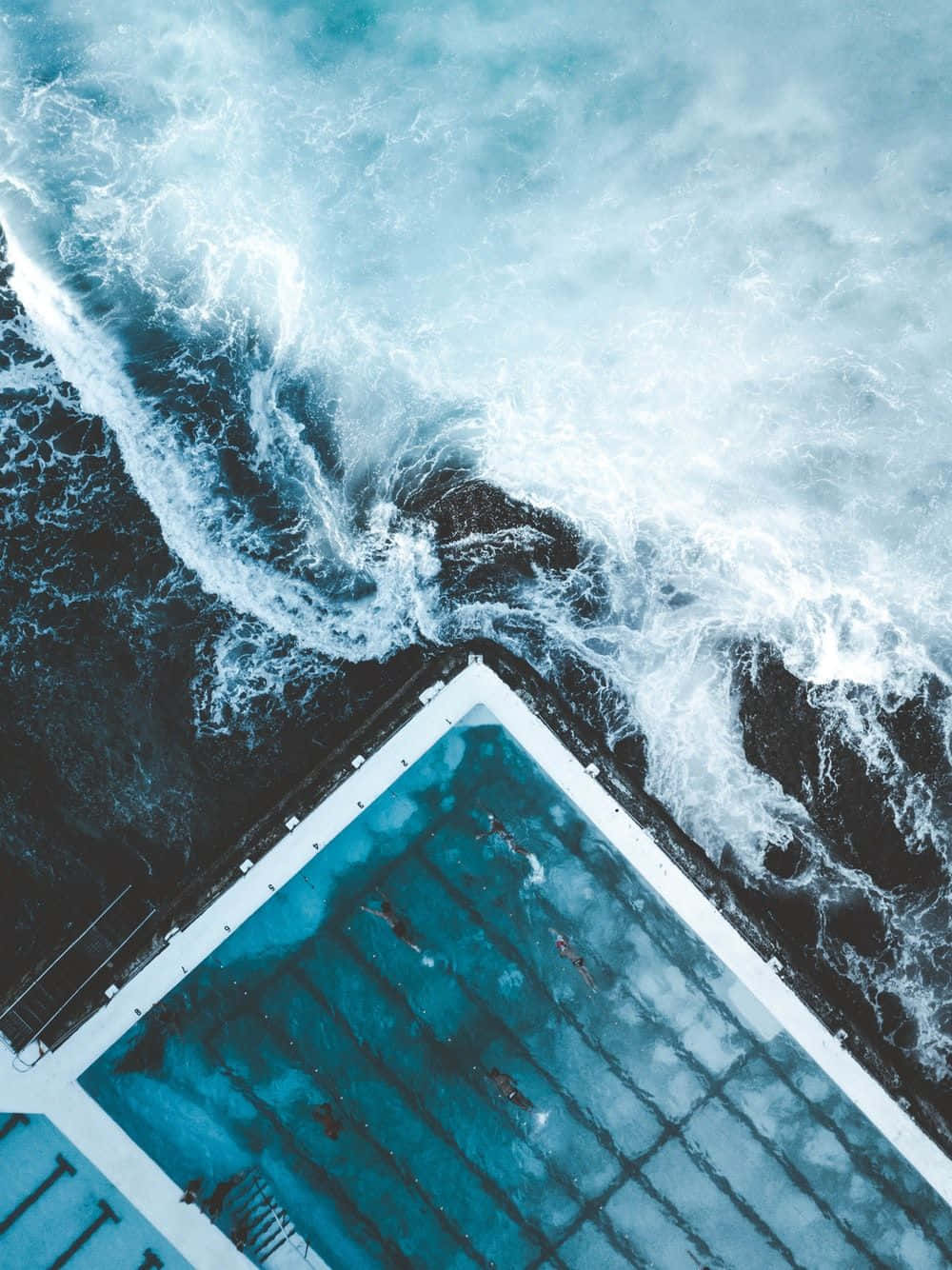Swimming Pool iPhone 7 Plus Live 壁紙 — 俯瞰海洋波浪撞擊幾何形游泳池與游泳者的景象。
