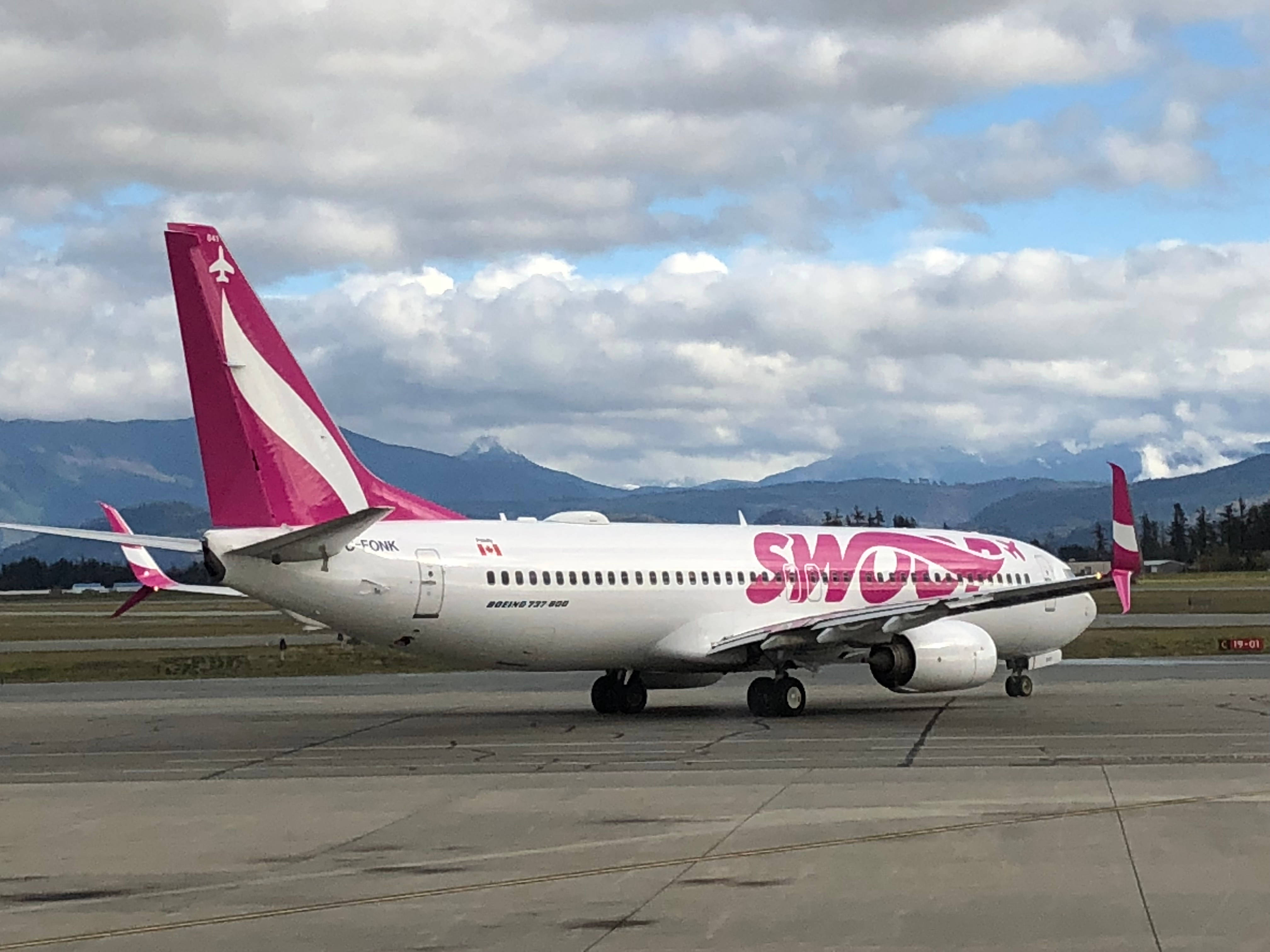 Swoop航空準備起飛 壁紙 — 一架粉紅色和白色的Swoop Airlines波音737停泊在停機坪上，背景是白雪覆蓋的山脈和多雲的天空。