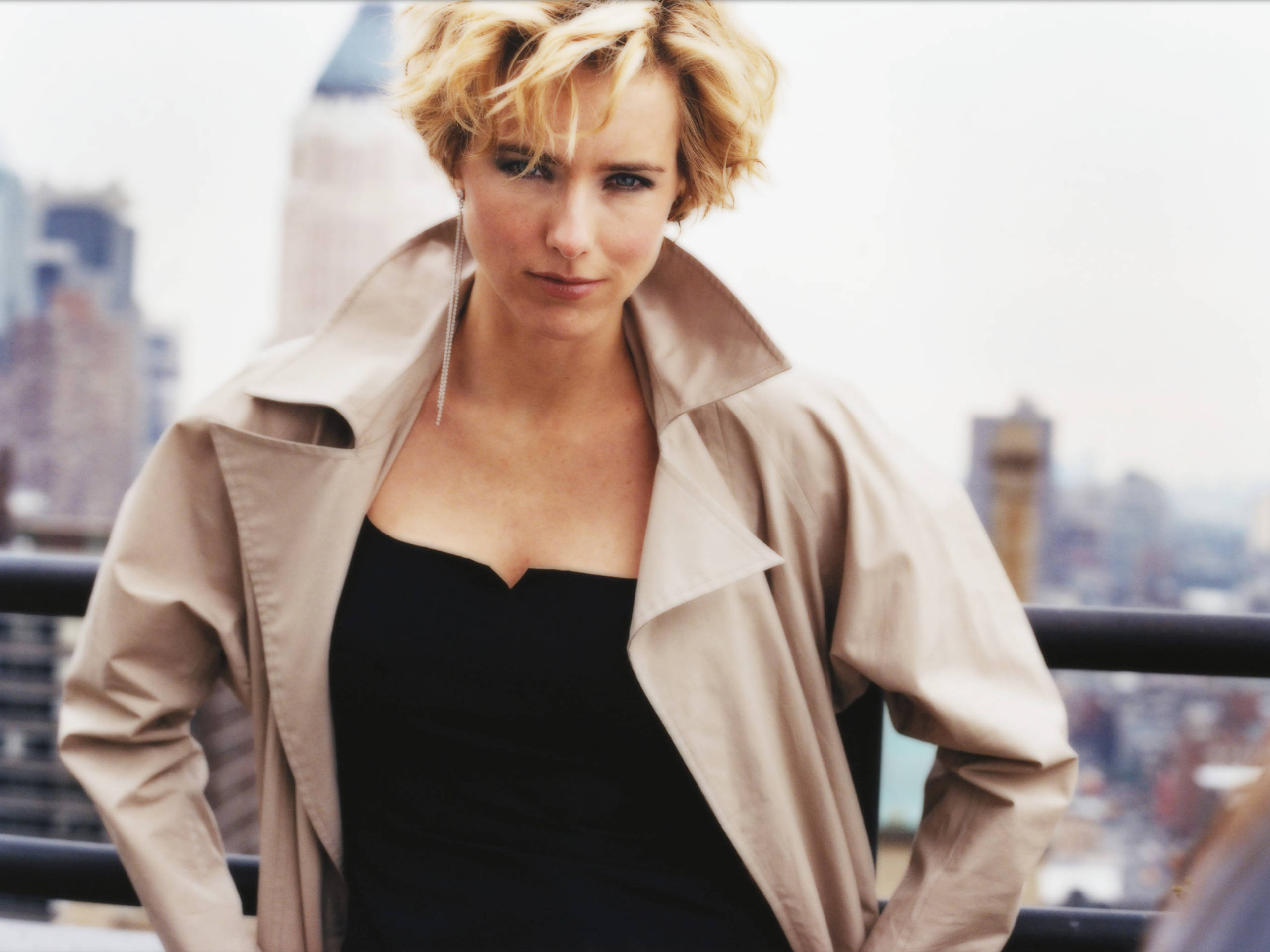 Téa Leoni 米色外套 壁紙 — 一位短金髮女性穿著米色風衣，自信地站在城市屋頂上，俯瞰模糊的城市天際線。