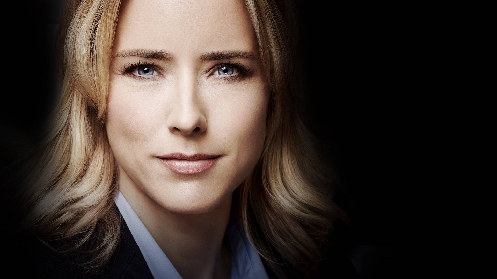 Téa Leoni 黑底背景 壁紙 — 一位金髮藍眼女士穿著深色西裝，在黑色背景前的特寫肖像。