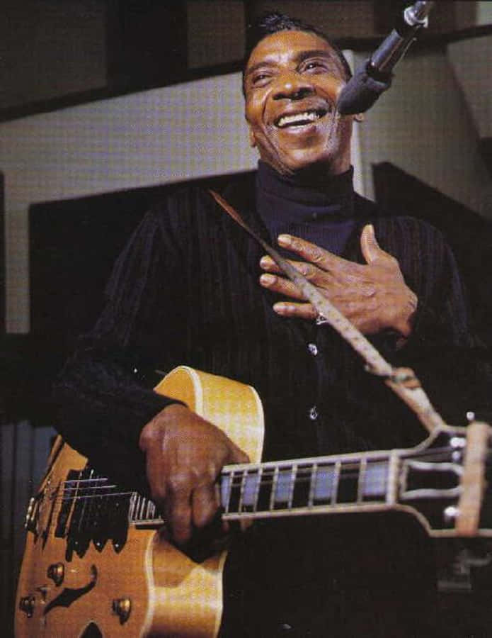 最偉大的藝術家 — T-Bone Walker 壁紙 — 一位快樂的音樂家在溫暖的舞台燈光下，一邊彈奏著旭日紋電結他，一邊露出燦爛的笑容。