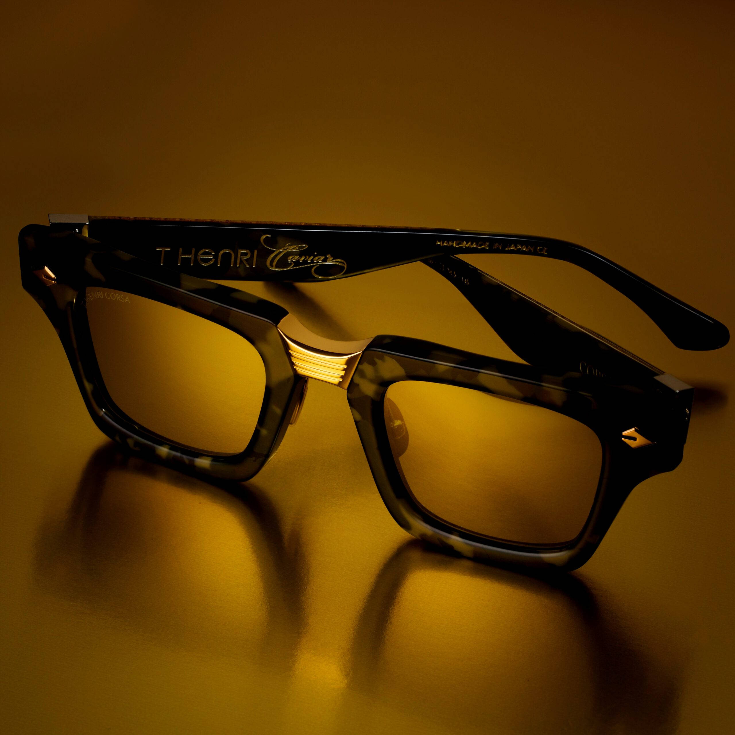 T Henri Caviar眼鏡 壁紙 — 一副帶有金色點綴的玳瑁太陽眼鏡，在柔和的方向性燈光下，放置在溫暖的金色表面上。