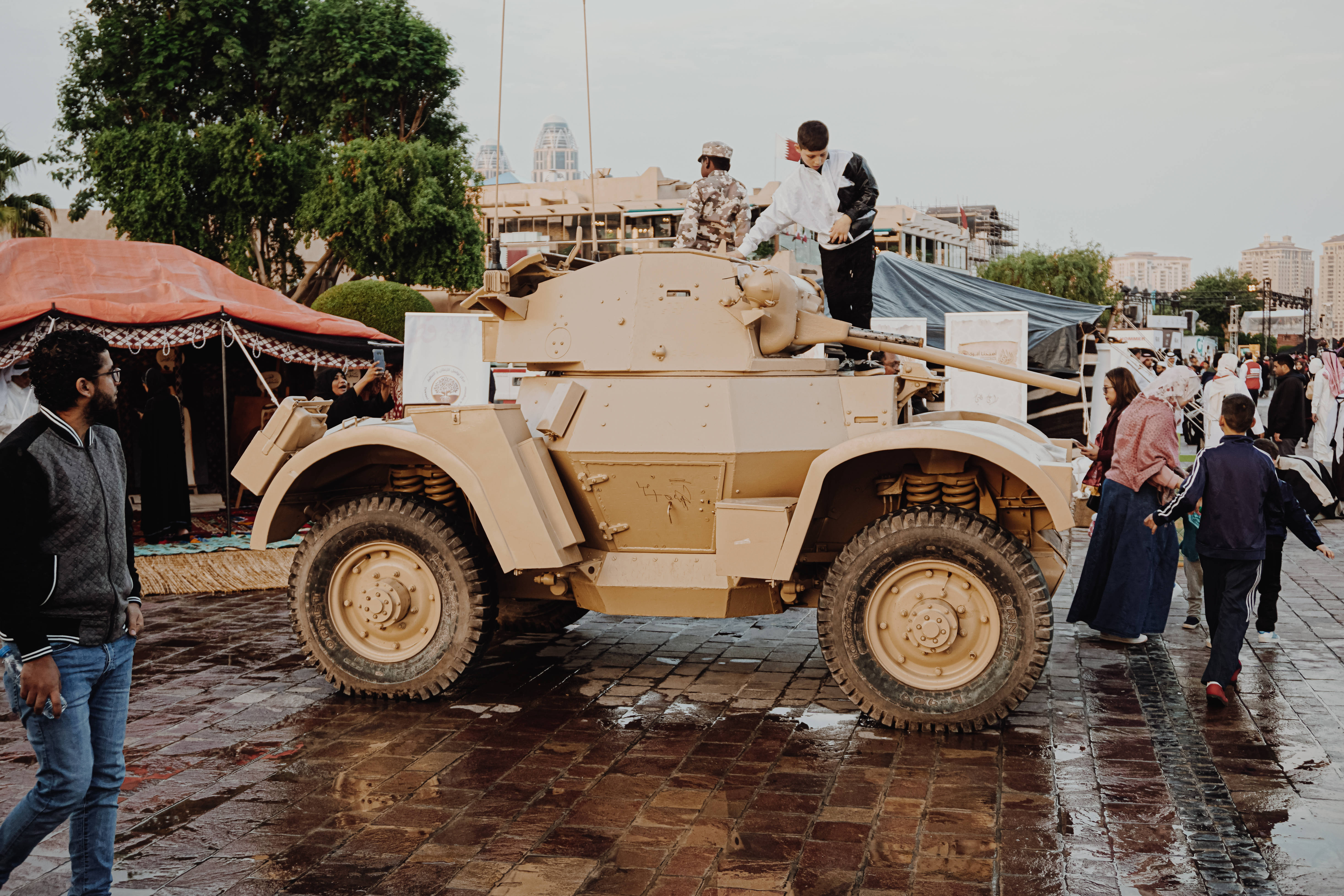 T17E1 Staghound 坦克模型 壁紙 — 一輛米色裝甲車配備大型輪胎停在濕滑的鵝卵石上，人們在附近走動，孩子們在陰天玩耍。