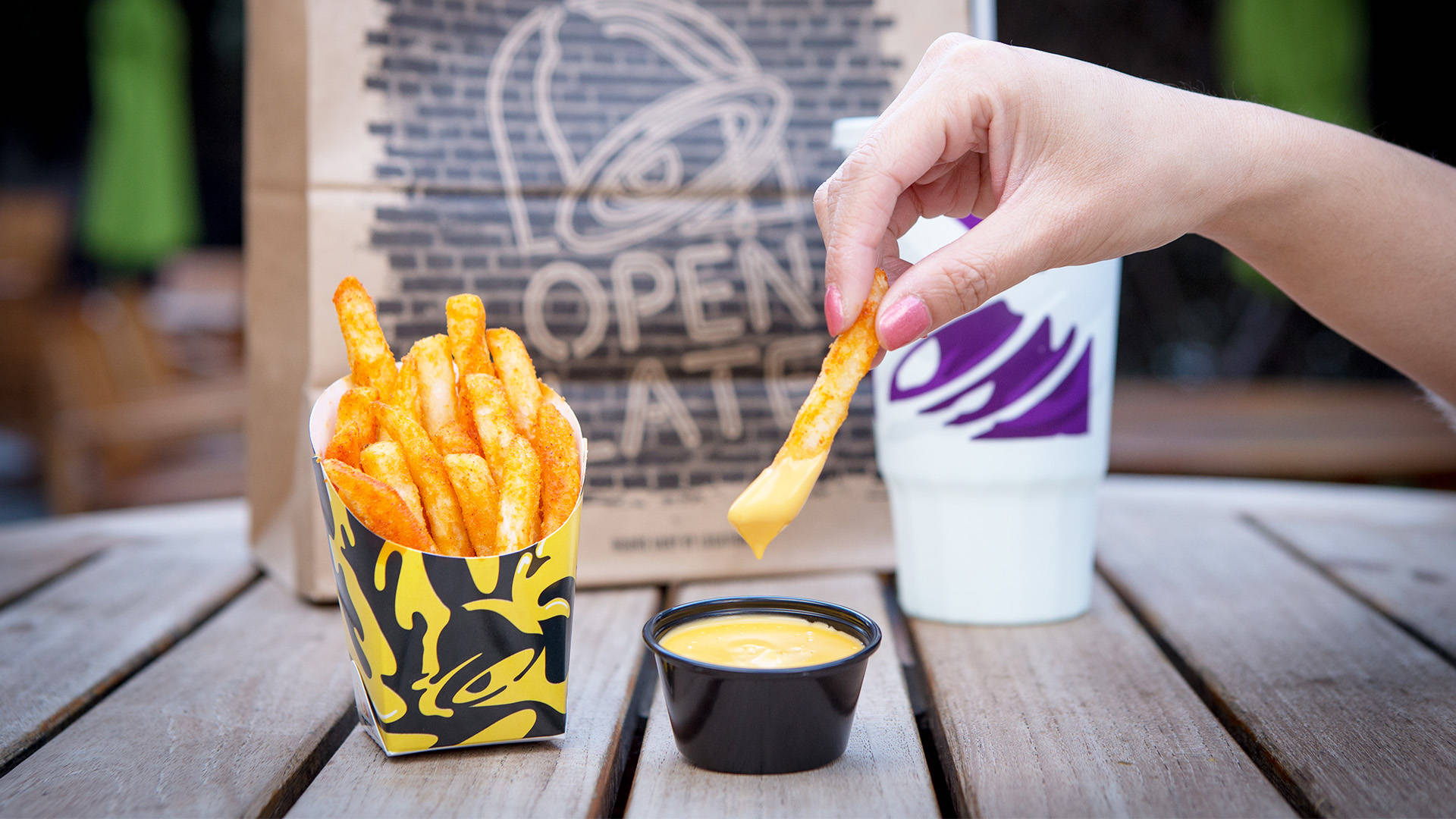 Taco Bell Nacho薯條與芝士醬 壁紙 — 一隻手將辣薯條蘸入芝士醬中，旁邊有一個品牌杯和盒子放在木桌上。