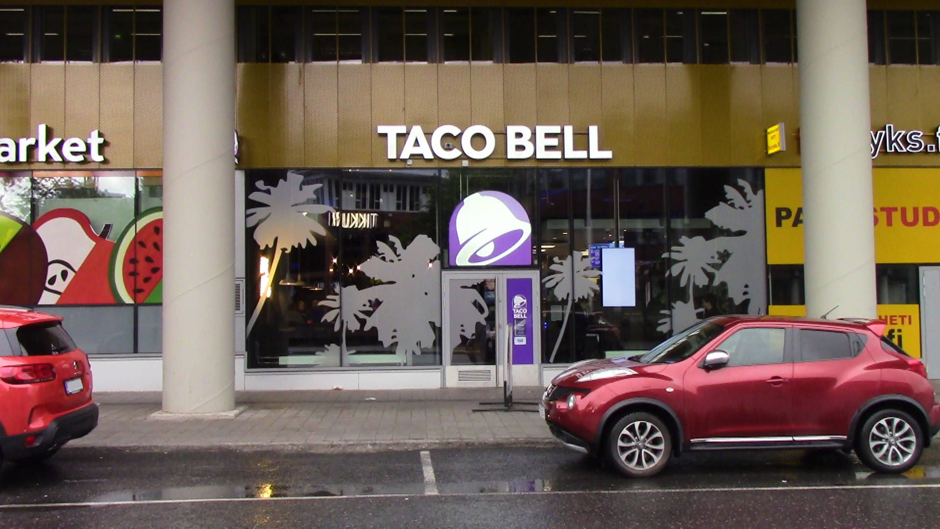 馬來西亞迪克西高速公路旁的 Taco Bell 壁紙 — 一輛紅色SUV停在Taco Bell餐廳前，玻璃窗上有棕櫚樹貼紙，濕漉漉的人行道反射著城市燈光。