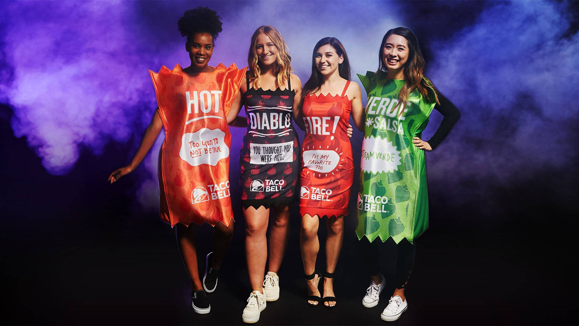 Taco Bell萬聖節服裝 壁紙 — 四位微笑的女性穿著Taco Bell醬料服裝，在煙霧繚繞的紫色和藍色背景前擺姿勢。