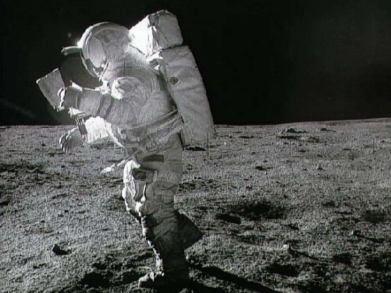 Astronaut On Moon 桌布 壁紙 — 一名穿著笨重太空衣的太空人在黑色天空下走過月球荒蕪的灰色表面。