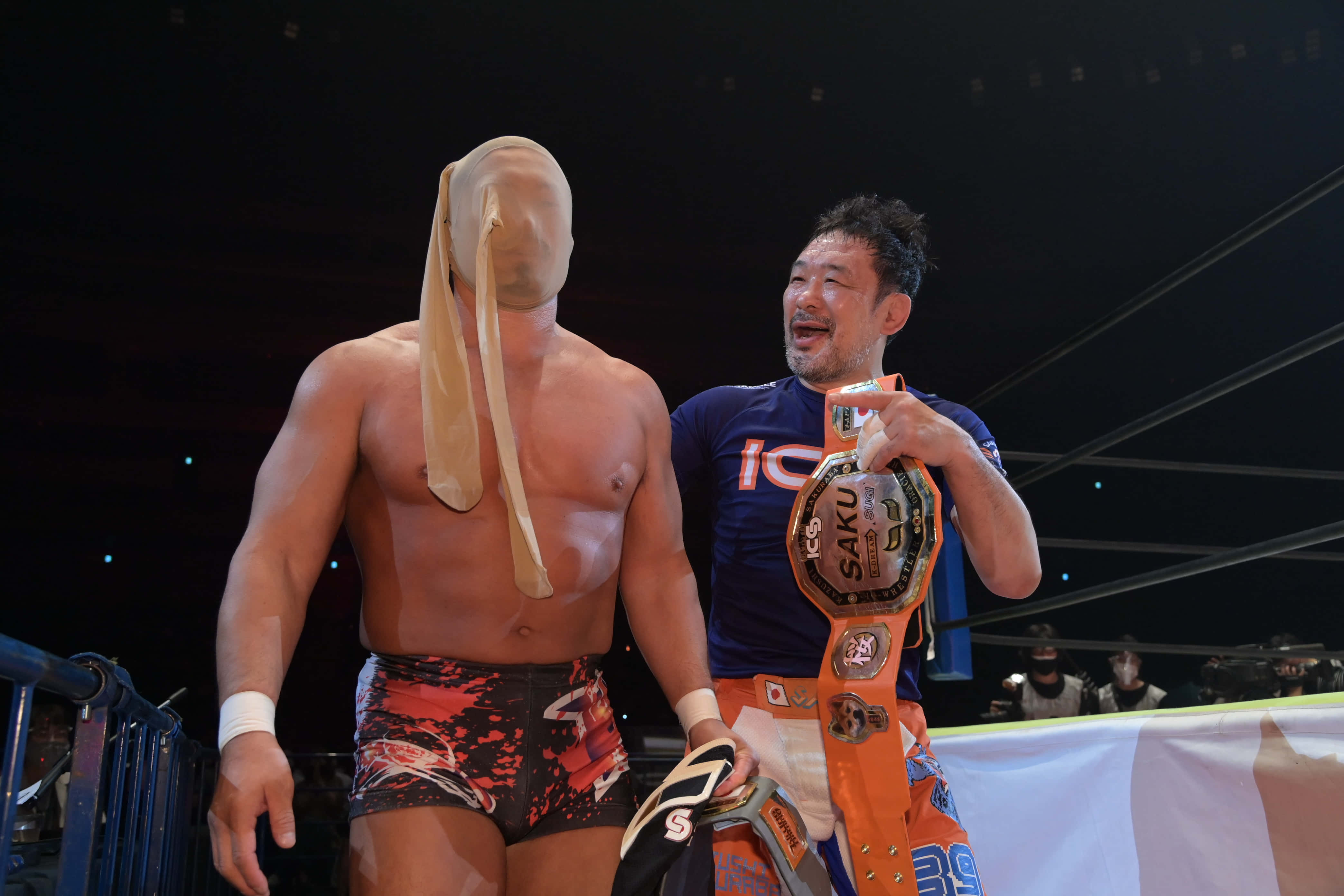 Takashi Sugiura 與 Kazushi Sakuraba 壁紙 — 兩名摔角手在擂台上，一人戴著面具，另一人手持冠軍腰帶，身穿亮橙色裝備，在場館燈光下。
