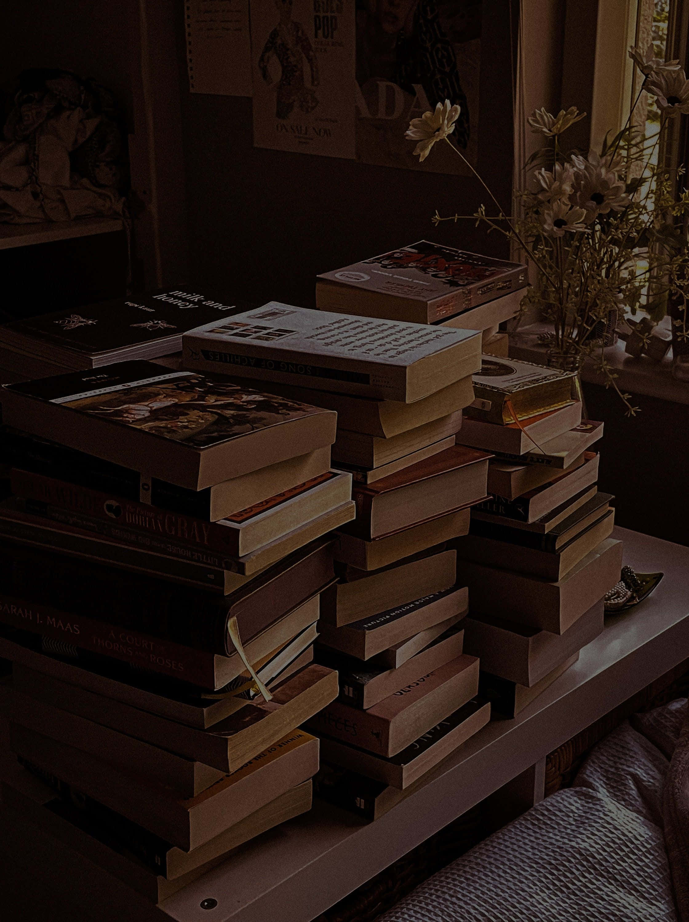Dark Book Aesthetic 桌布 壁紙 — 高高的書堆放在木桌上，旁邊是窗戶，窗戶上有乾花和海报，房間光線昏暗。