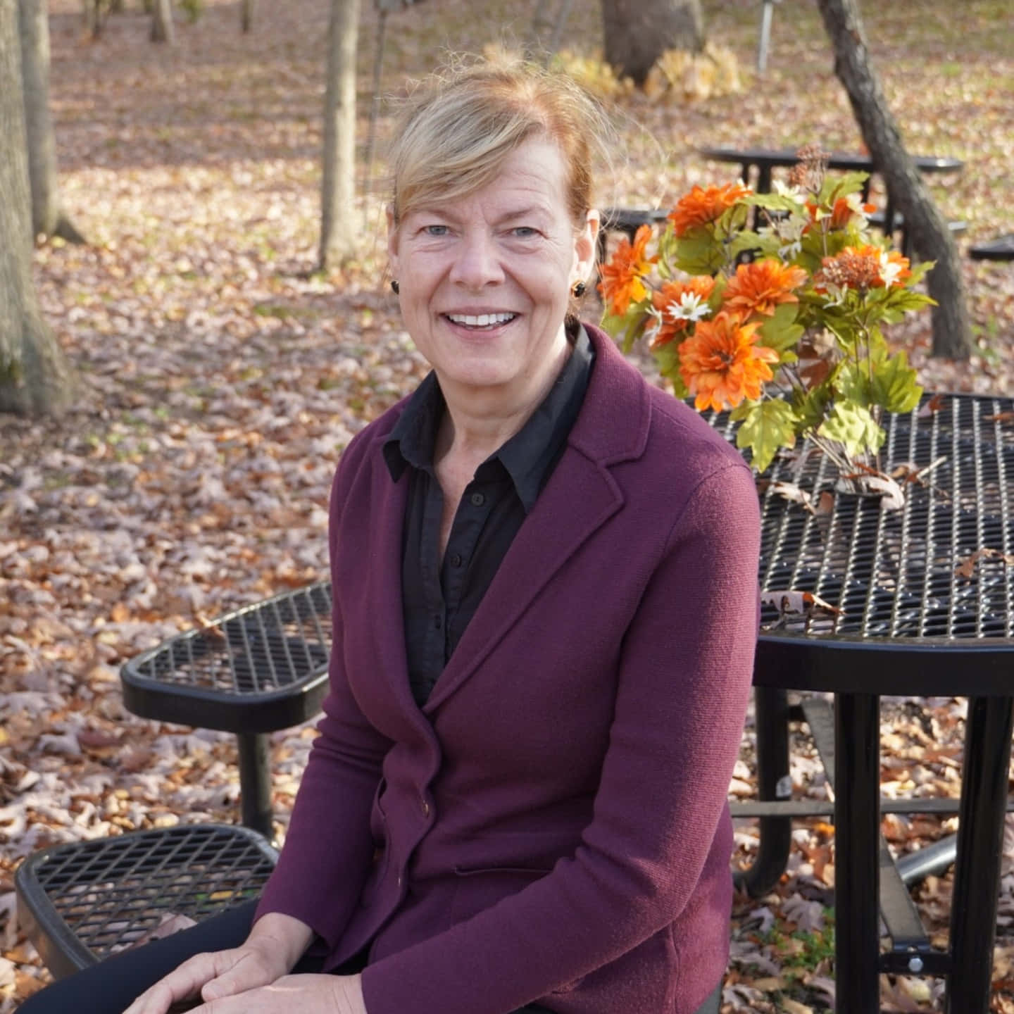 Tammy Baldwin 坐在金屬長椅上 壁紙 — 一位微笑的女子穿著紫色西裝外套，坐在金屬野餐桌旁，周圍是秋天的落葉和鮮豔的橙色花朵。