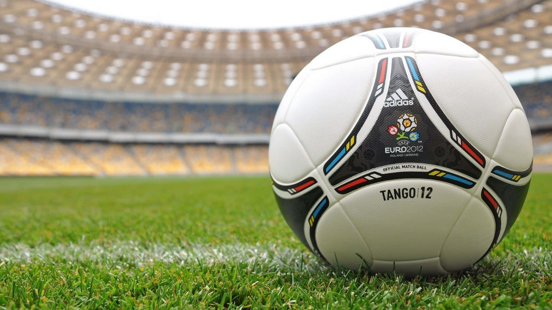 Tango 12足球背景 壁紙 — 一個白色的 Adidas Tango 12 足球放在一個空蕩蕩的體育館的鮮綠色草地上，體育館有分層座位。