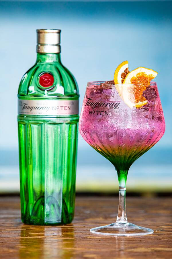 Tanqueray No. Ten金酒與粉紅雞尾酒 壁紙 — 一杯充滿活力的粉紅色氈酒雞尾酒配以橙裝飾，放在海邊木桌上一支綠色Tanqueray酒瓶旁邊。