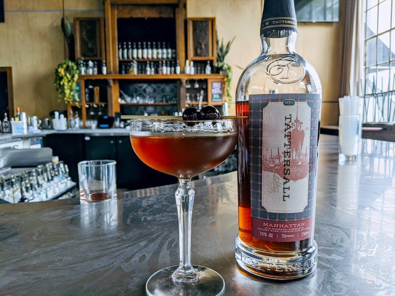 Tattersall Distilling 曼哈頓雞尾酒 壁紙 — 一杯豐富的琥珀色曼哈頓雞尾酒，飾以櫻桃，放在其酒瓶旁邊，在溫暖燈光的酒廊中拋光吧檯上。