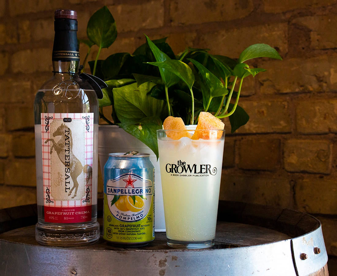 Tattersall Distilling 精緻手工雞尾酒 壁紙 — 一杯清爽的西柚雞尾酒，飾以橙片，旁邊放著一瓶Tattersall西柚忌廉和San Pellegrino柚子，放在木桶上，背景是磚牆和綠色植物。