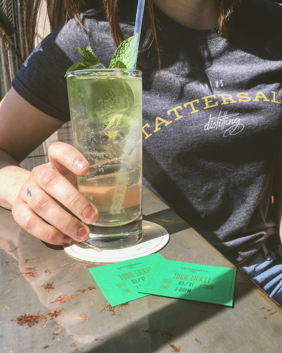 於 Tattersall Distilling 享用清爽莫吉托 壁紙 — 一個人在明媚陽光下手持薄荷雞尾酒，綠色旅遊門票放在一張風化的桌子上。