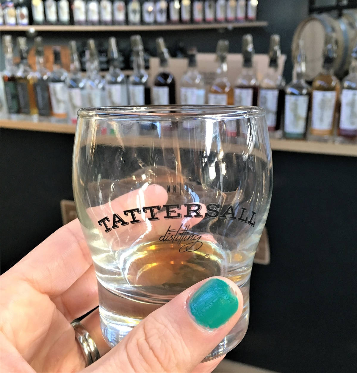 Tattersall 蒸餾廠特色烈酒杯 壁紙 — 一隻塗有青綠色指甲油的手，握著一個Tattersall Distilling玻璃杯，內含琥珀色液體，背景是威士忌酒瓶。