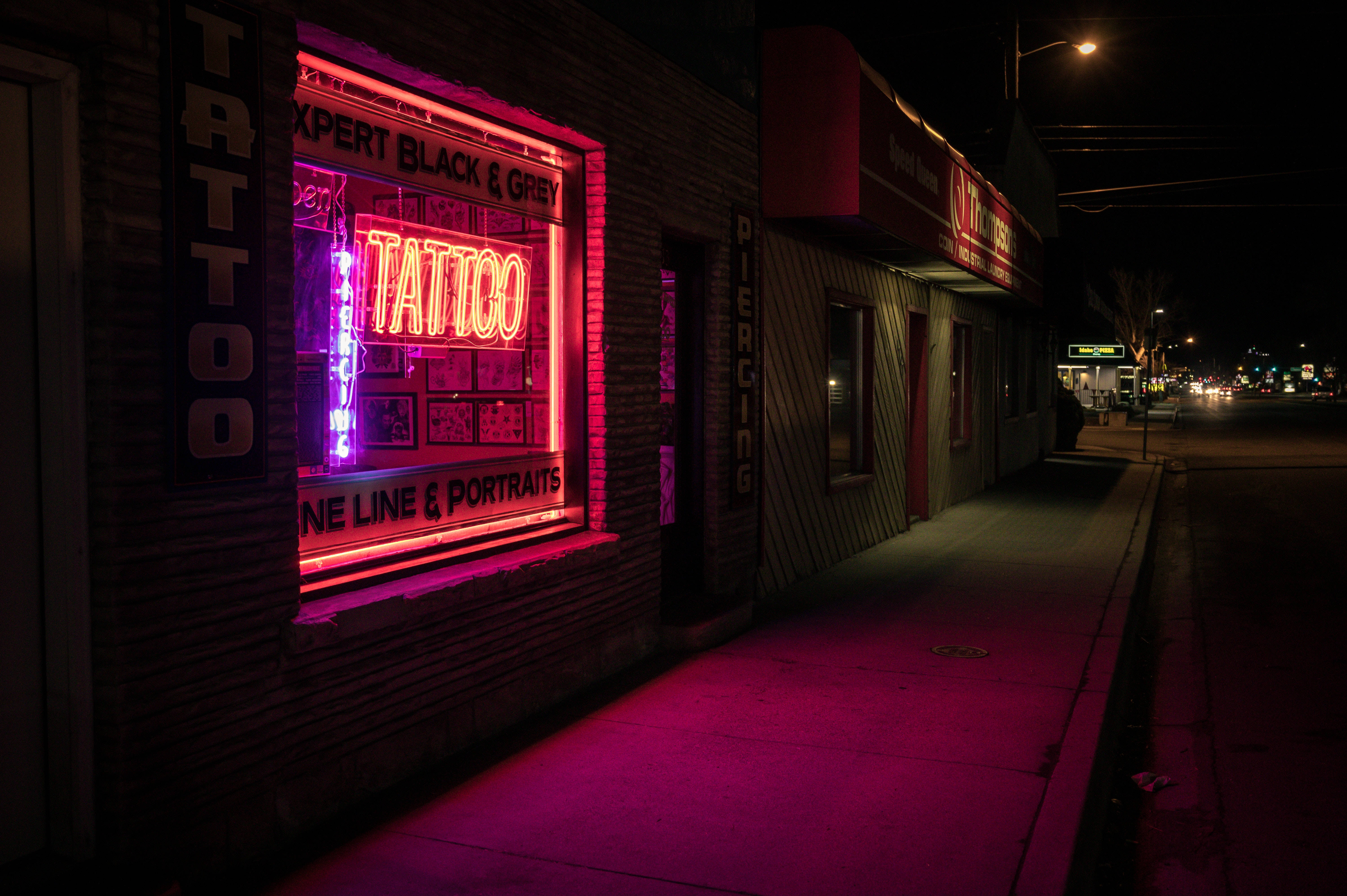 窗戶上的「TATTOO」霓虹燈招牌 壁紙 — 一個充滿活力的粉紅色霓虹紋身店招牌在漆黑的夜晚中發光，在人行道和磚牆上投射出超現實的光芒。