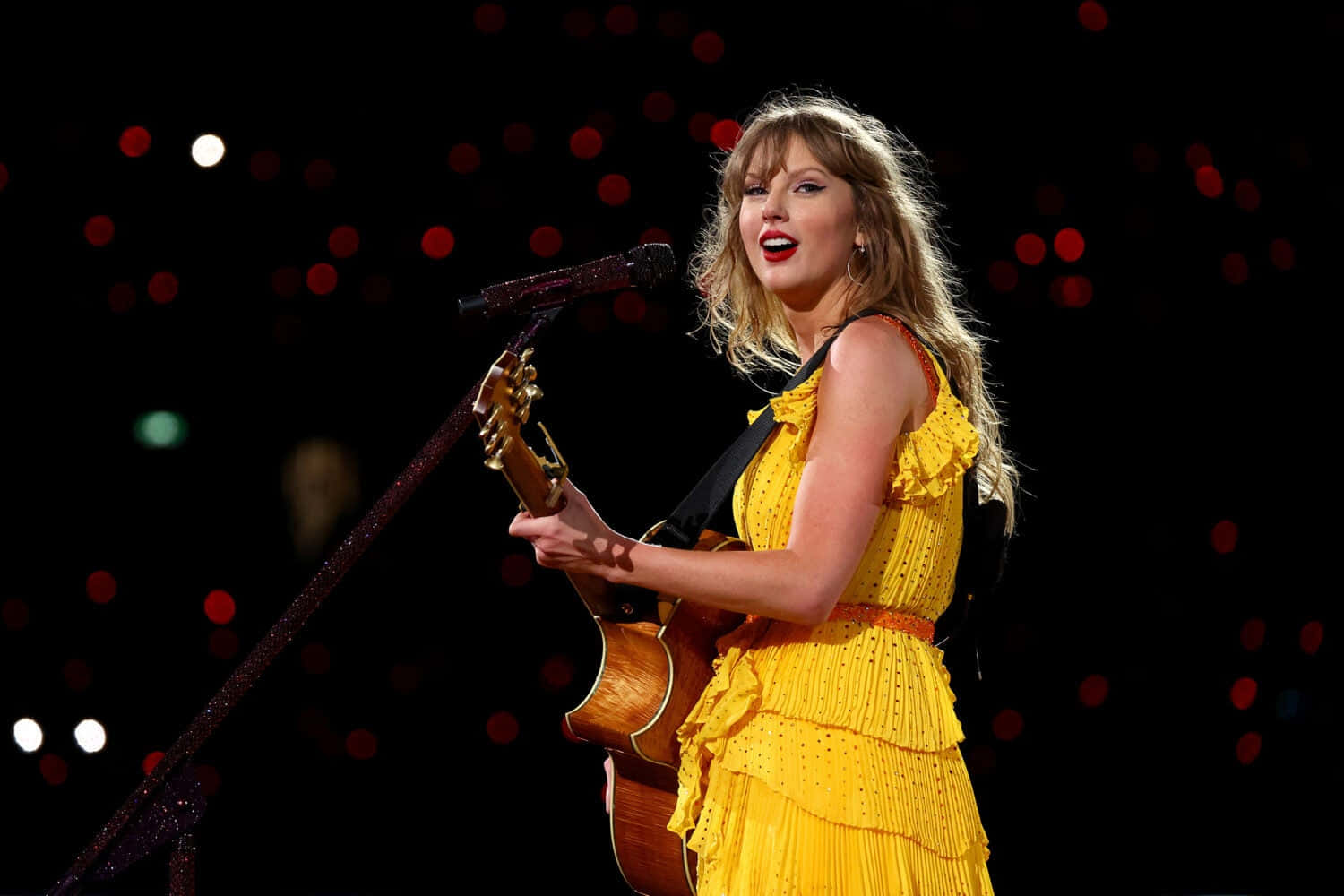  壁紙 — Taylor Swift 在舞台上穿著鮮黃色荷葉邊連衣裙，在聚光燈散景下拿著原聲結他表演。