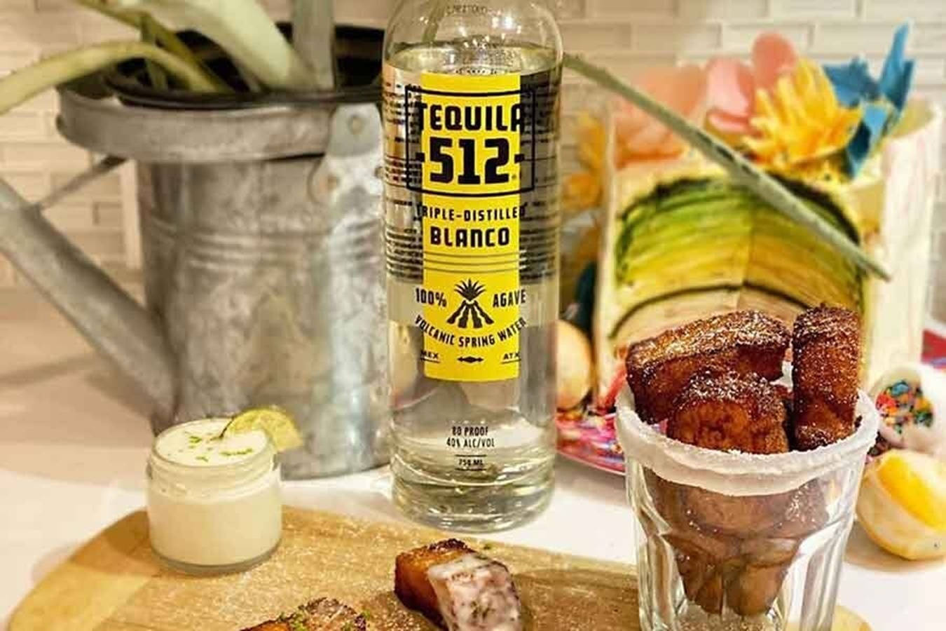 Tequila 512 Blanco 瓶身 壁紙 — 一瓶白龍舌蘭酒放在木板上，旁邊有脆豬皮和奶油蘸醬，背景有色彩繽紛的花朵和一個金屬桶。