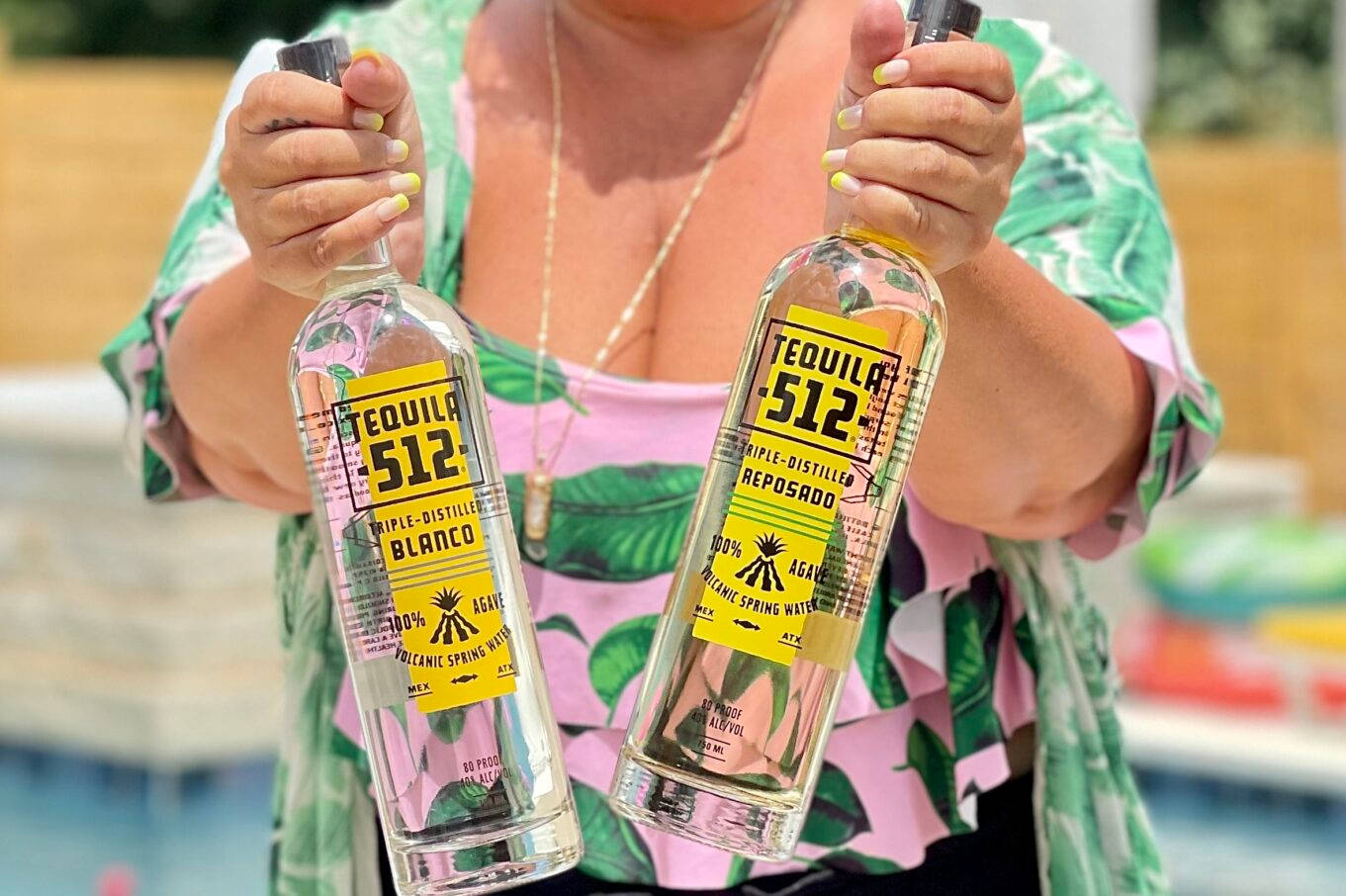 Tequila 512 Blanco 與 Reposado 瓶身由女士手持 壁紙 — 一位身穿熱帶印花上衣的女性在明媚陽光下，在泳池邊拿著兩瓶透明的Tequila 512。