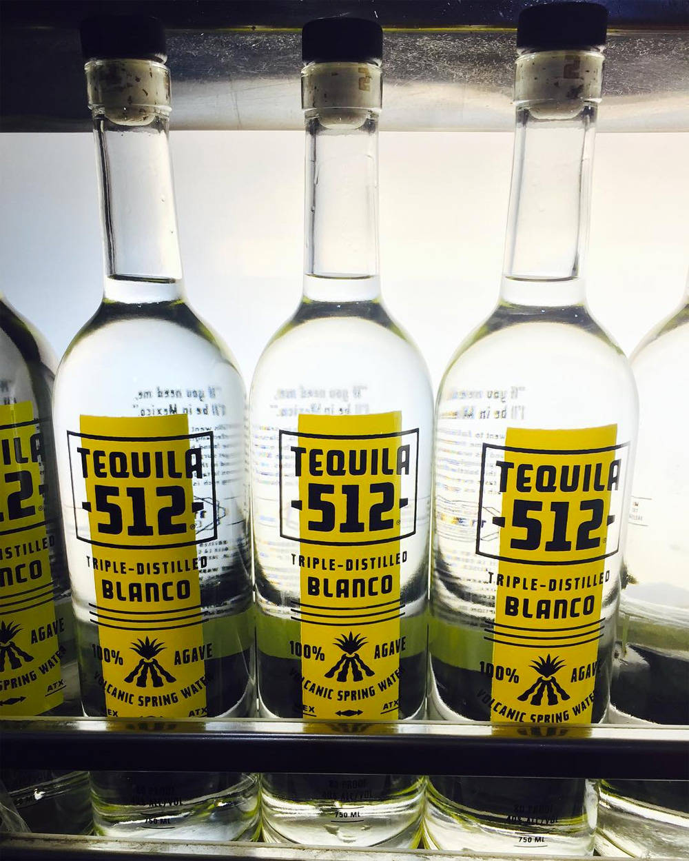 三瓶Tequila 512 白龍舌蘭酒 壁紙 — 一排排透明龍舌蘭酒瓶，帶有醒目的黃色標籤，整齊地擺放在明亮的燈光下的冷藏架上。