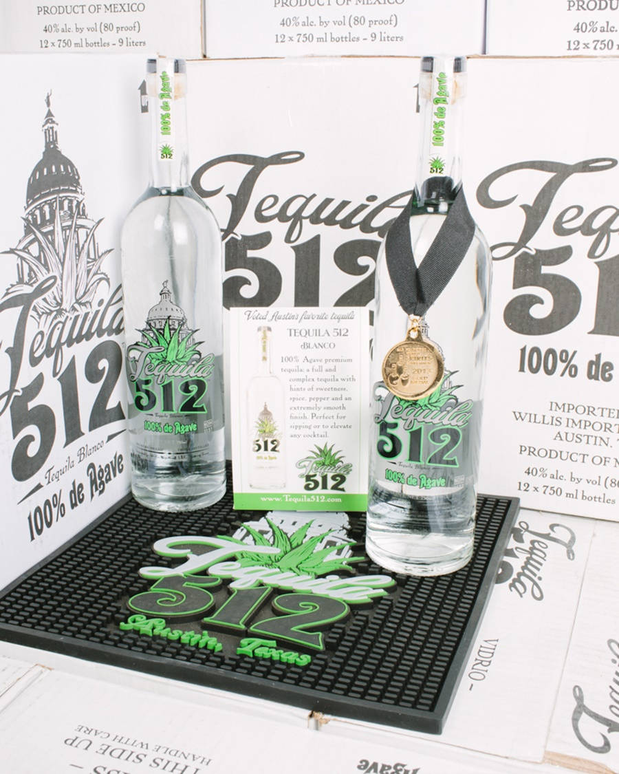 Tequila 512 白龍舌蘭酒透明瓶身 壁紙 — 兩支透明龍舌蘭酒瓶，帶有綠色標籤和一枚金牌，放在品牌墊上，背景是堆疊的產品盒。