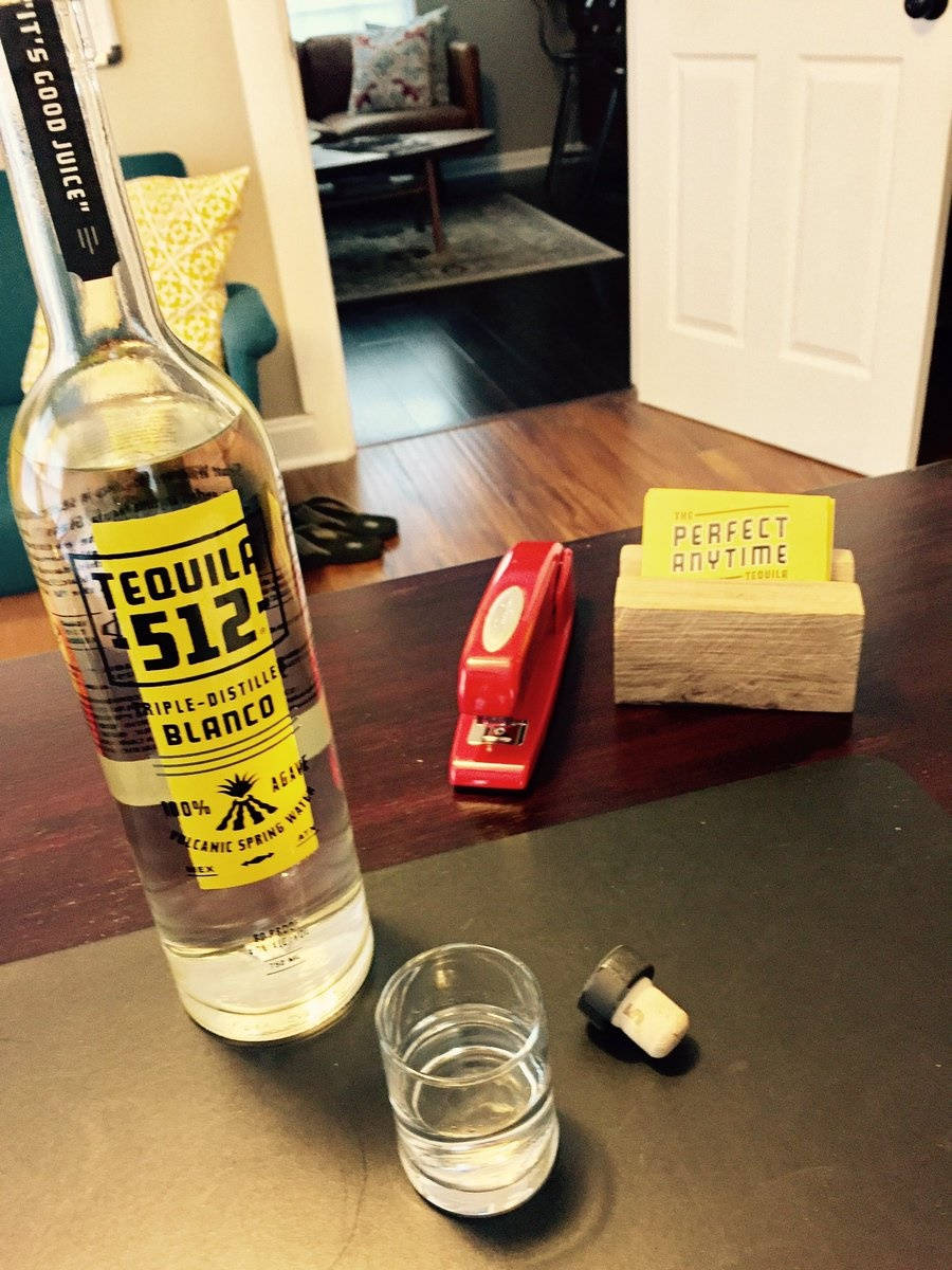 Tequila 512 白龍舌蘭酒室內實拍 壁紙 — 一個透明嘅龍舌蘭白瓶放在一張深色枱上，旁邊有一個杯同一個紅色釘書機連木製底座。