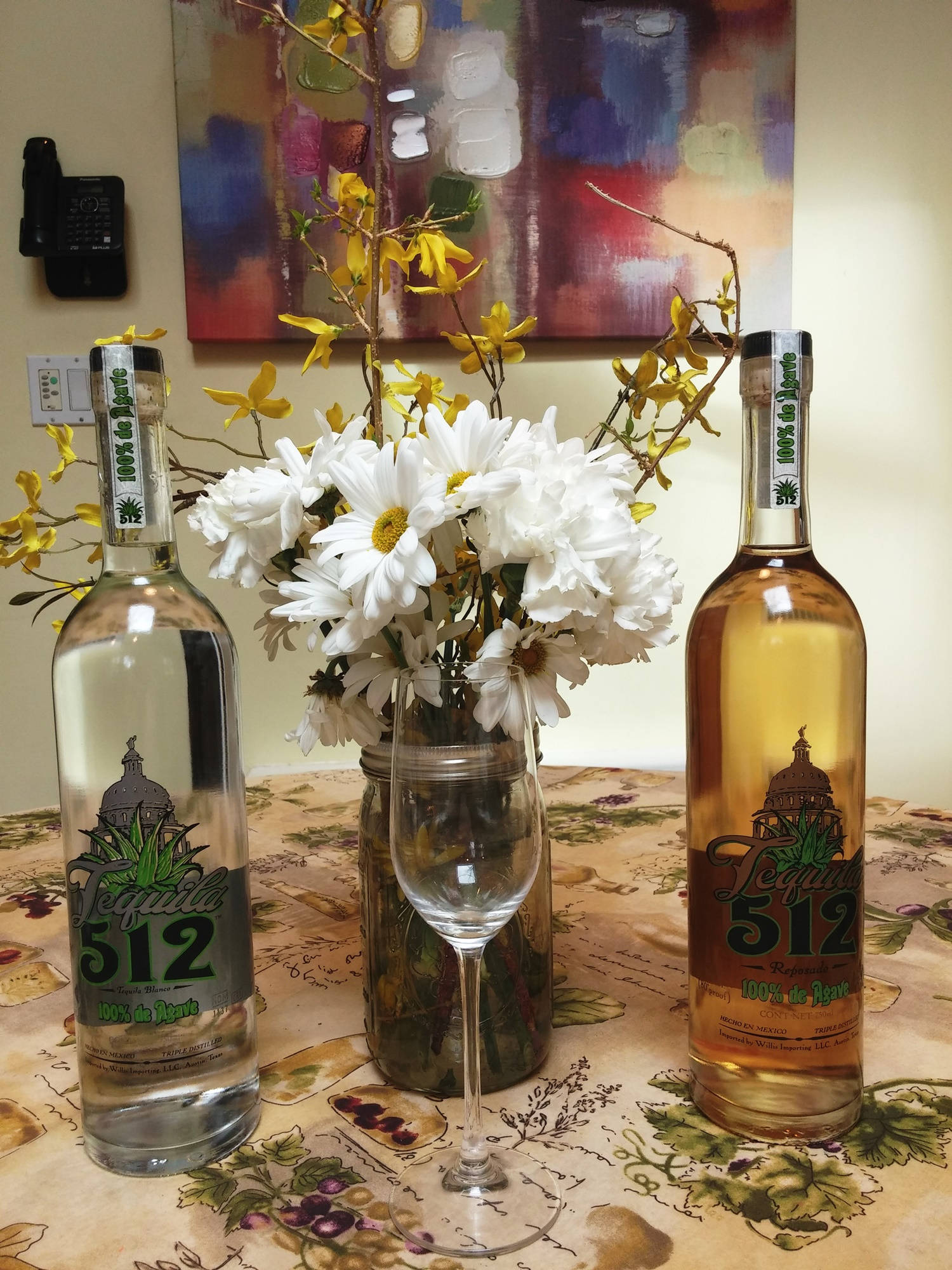 Tequila 512 灰黃標籤瓶身，展現傳統與創新的完美融合 壁紙 — 兩瓶龍舌蘭酒放在花桌布上，中間夾著一瓶白色雛菊和黃色連翹，在溫暖的室內燈光下。